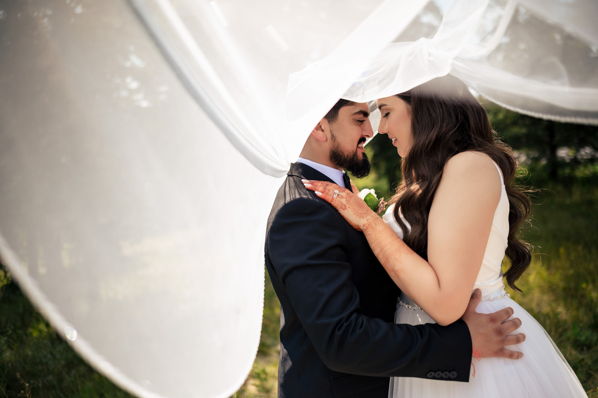 Bride and groom embrace under flowing veil outdoors.