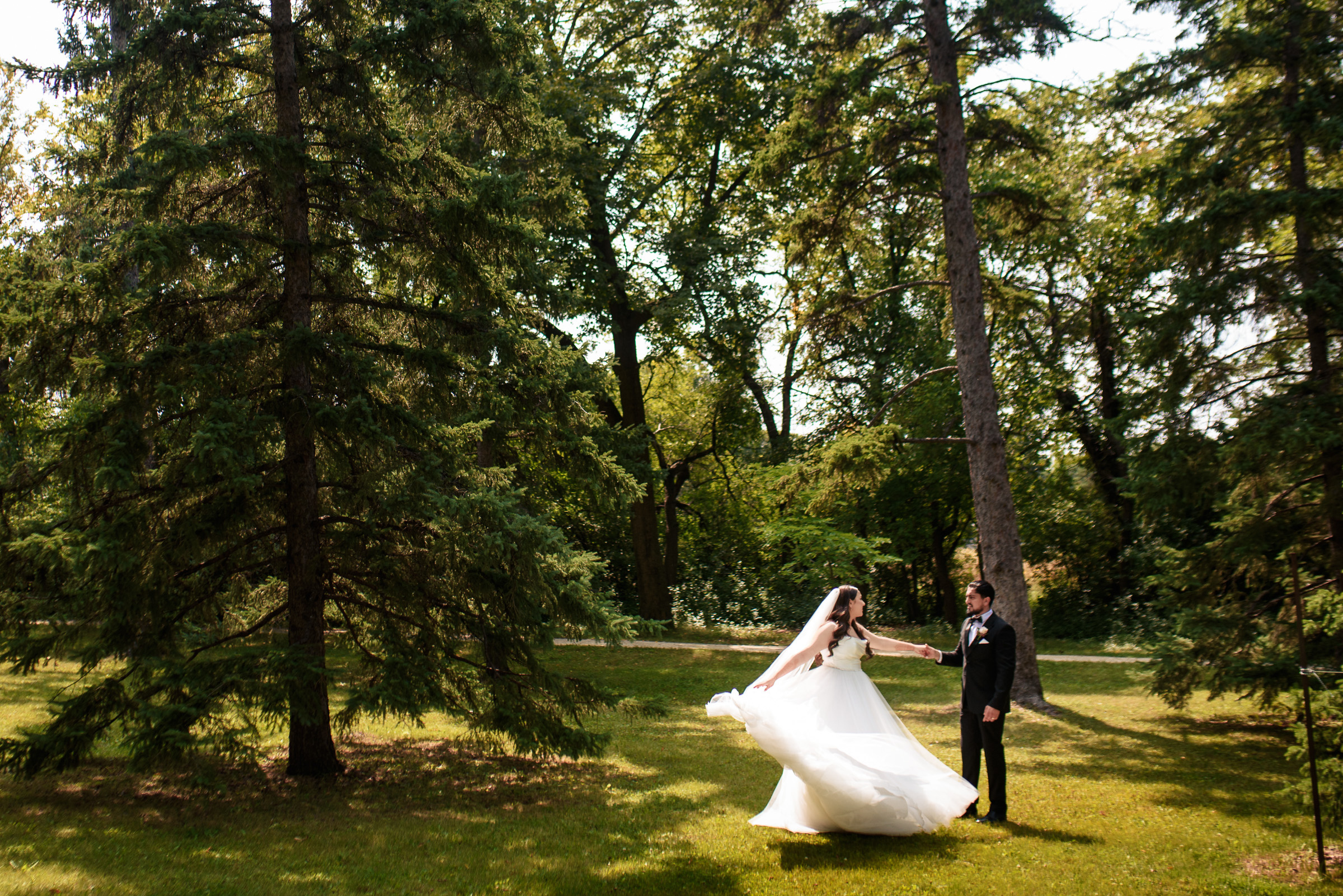 Bride twirling in wedding dress holding groom's hand in a sunlit forest setting.