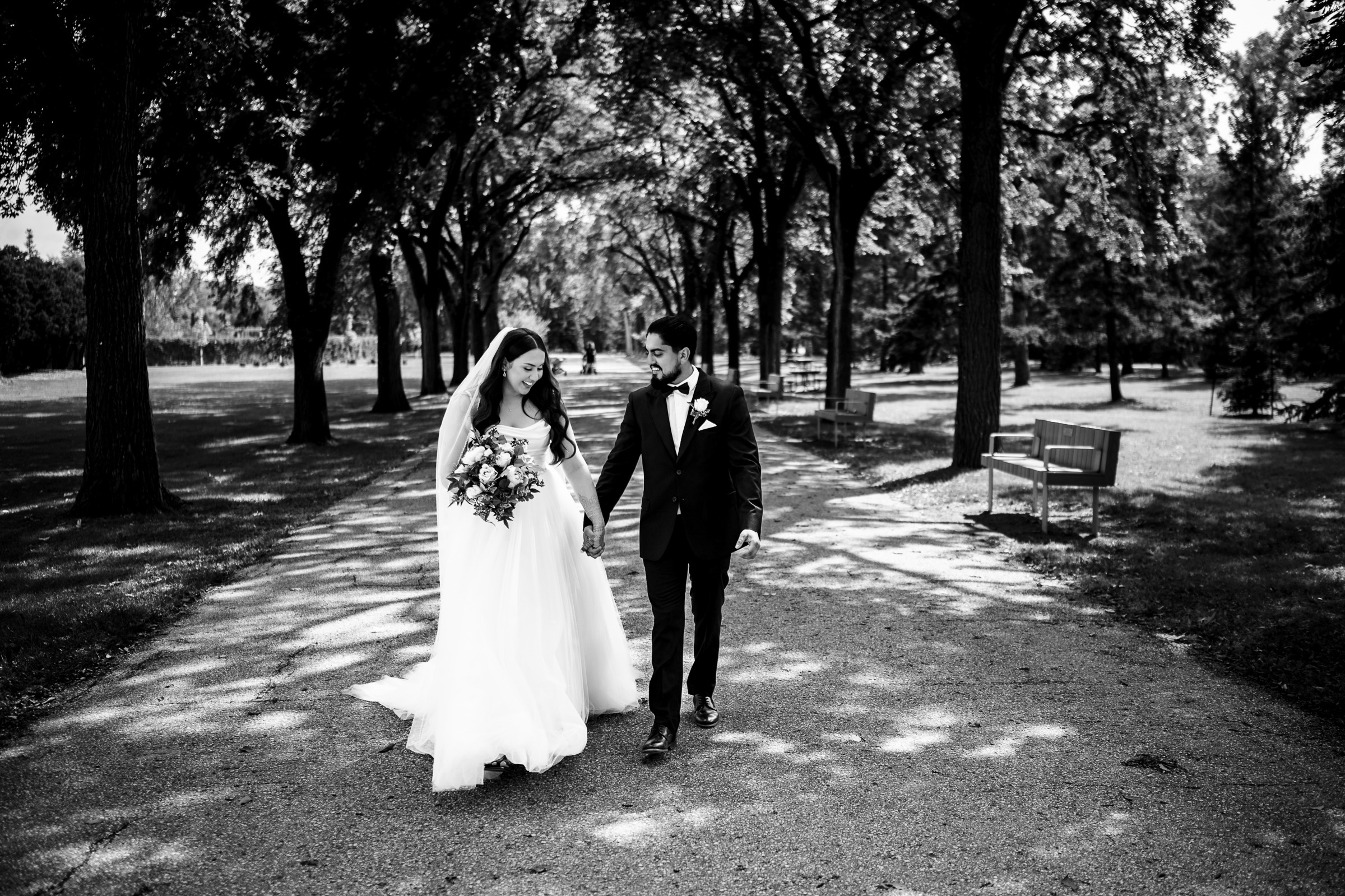 Bride and groom walking on a tree-lined path, holding hands and smiling.