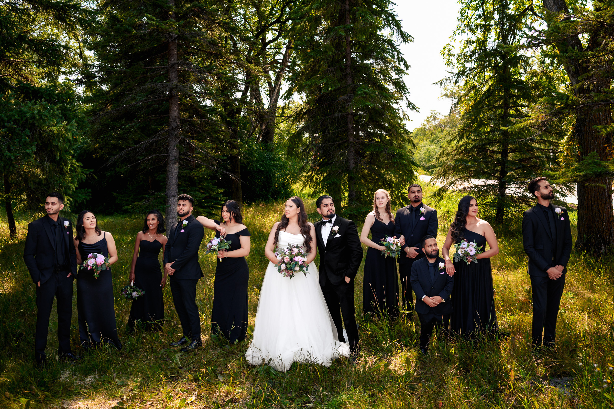 Bridal party stands in a forested area; bride in white, others in black attire with bouquets.