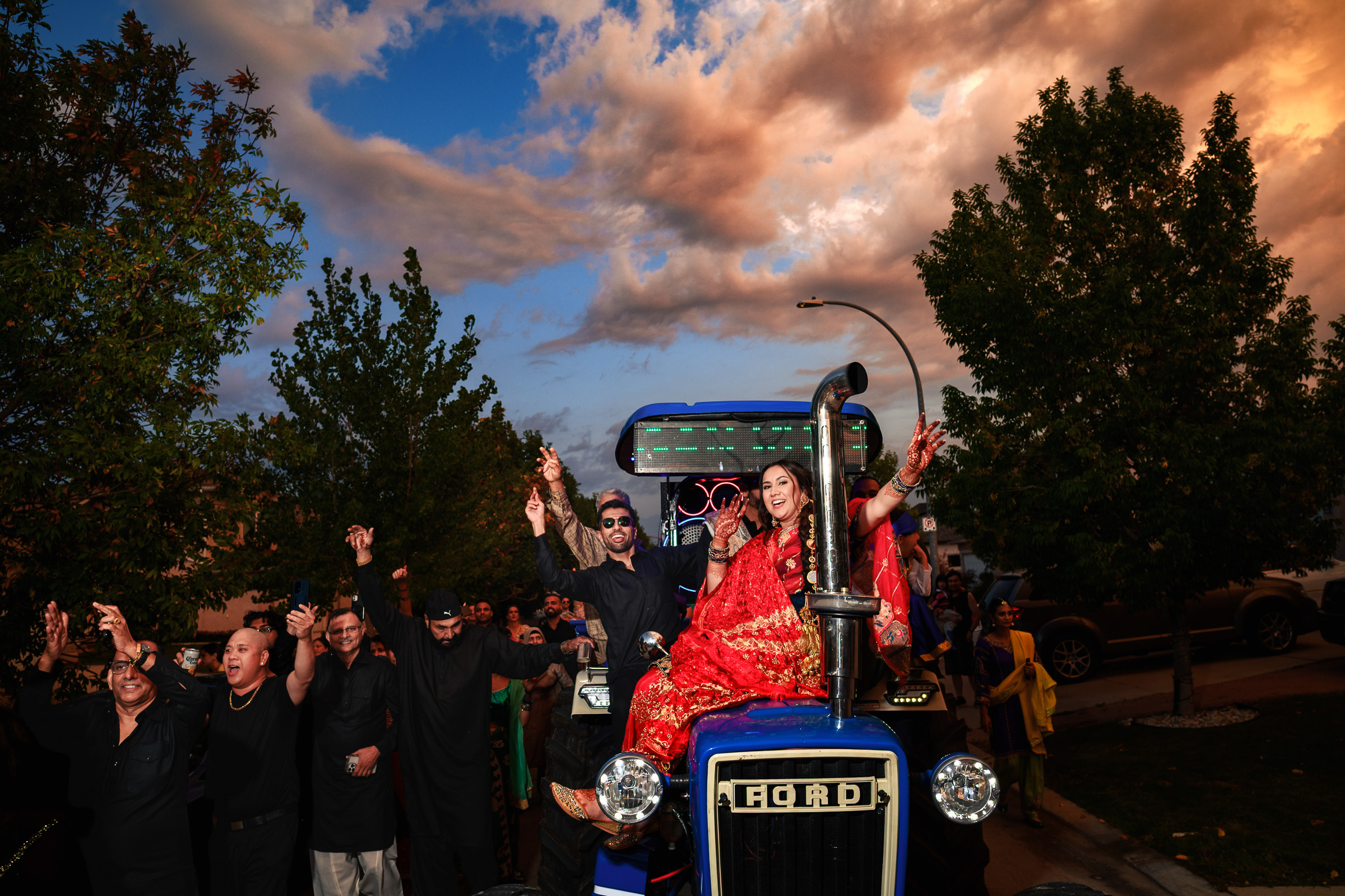 A couple in traditional attire rides a blue tractor, with people celebrating around them.