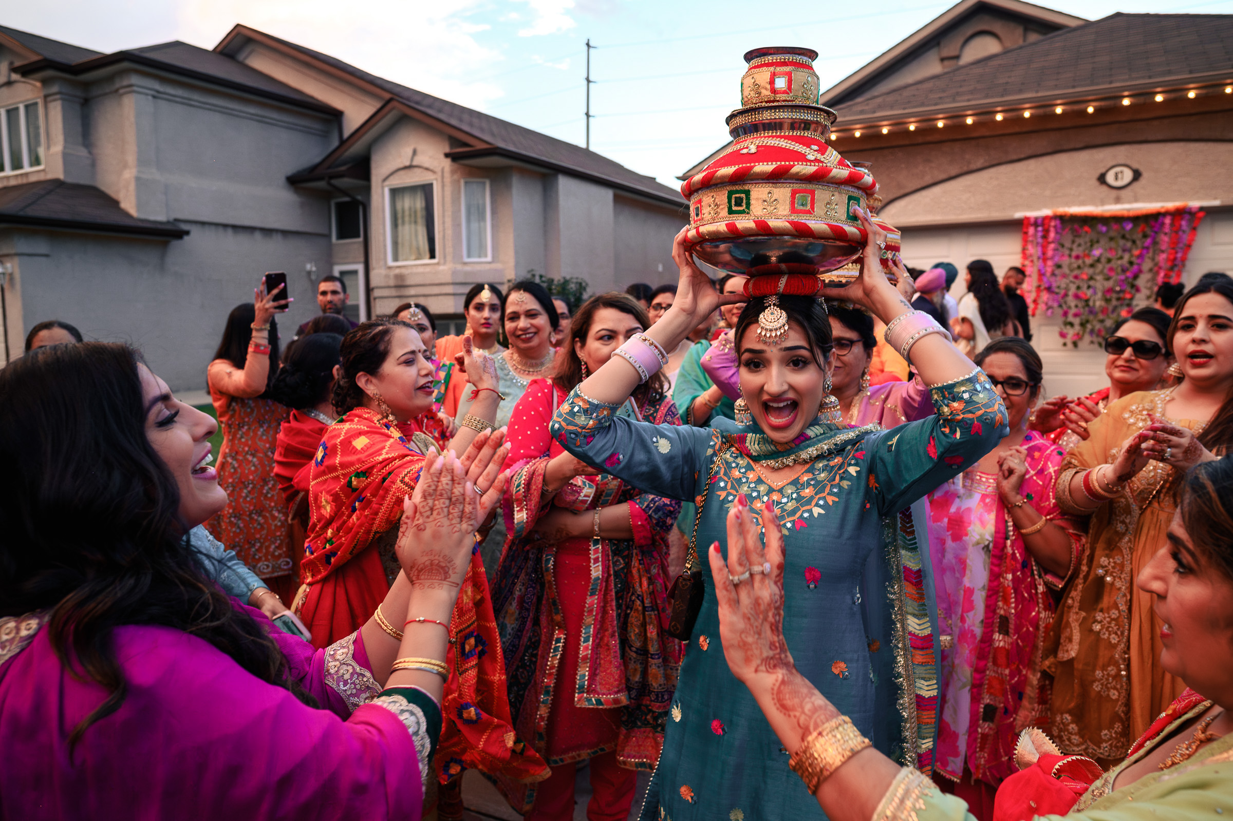 A woman in traditional attire holds a decorated pot overhead, surrounded by cheering people.