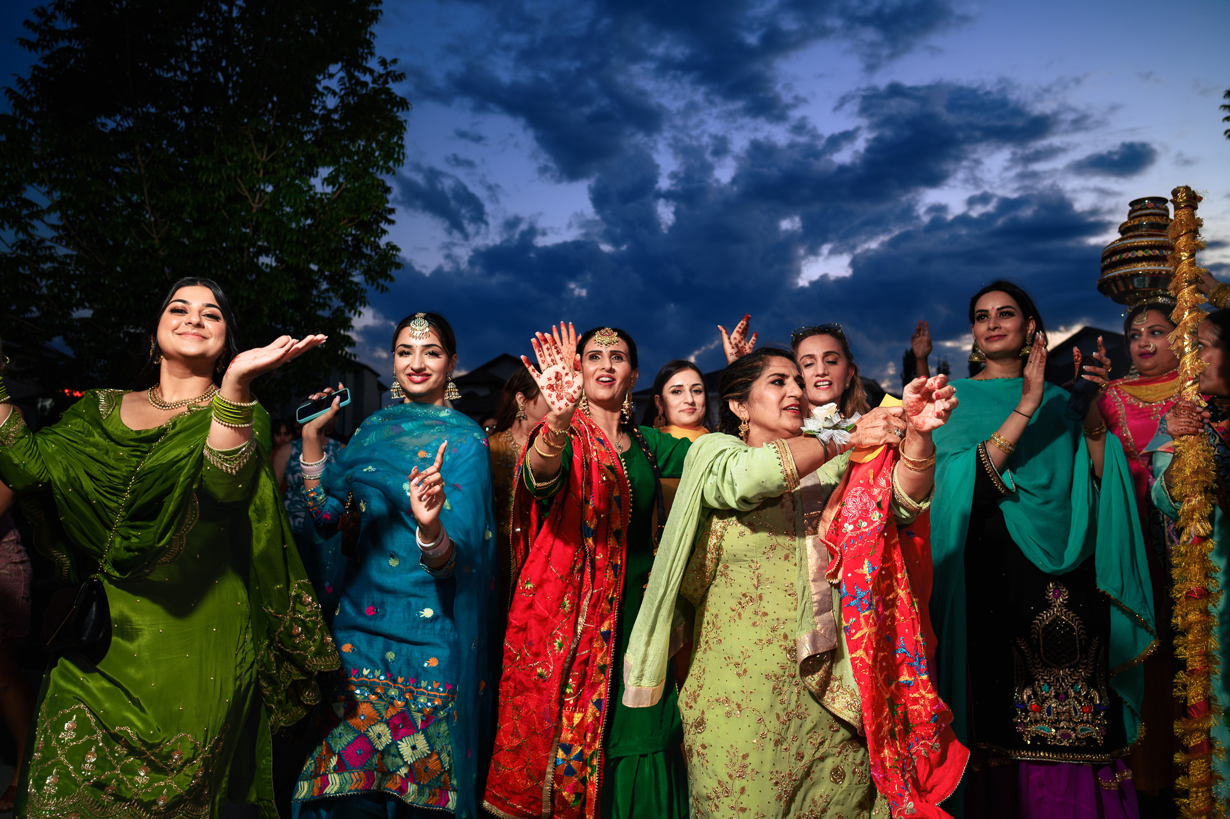 Women in vibrant traditional attire dancing outdoors under a cloudy evening sky.