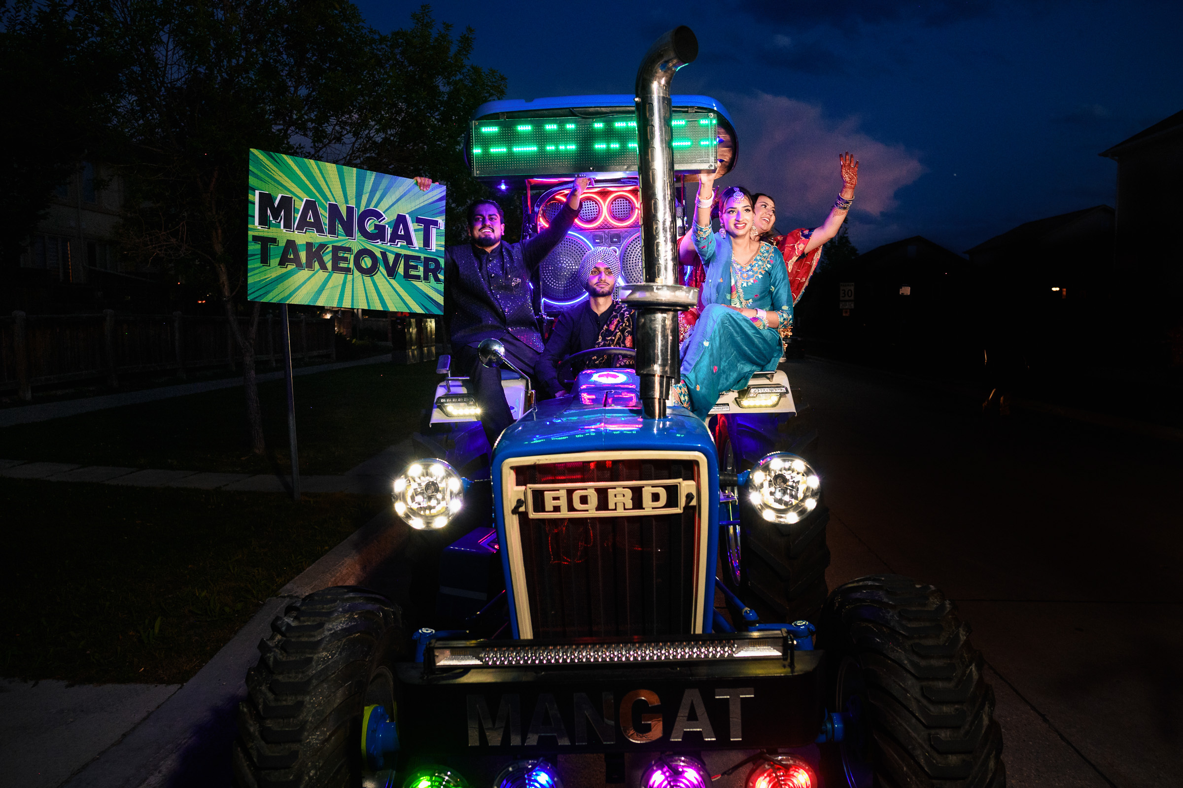Group posing on illuminated tractor with "Mangat Takeover" sign at night.