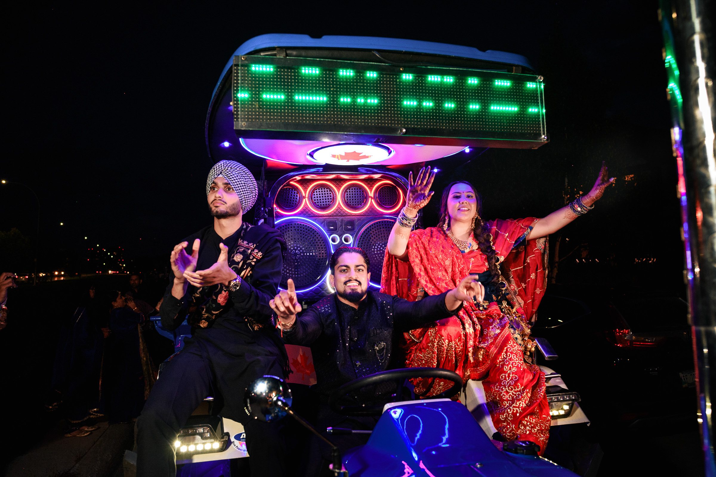 Three people in colorful attire ride a lit-up vehicle at night, celebrating energetically.