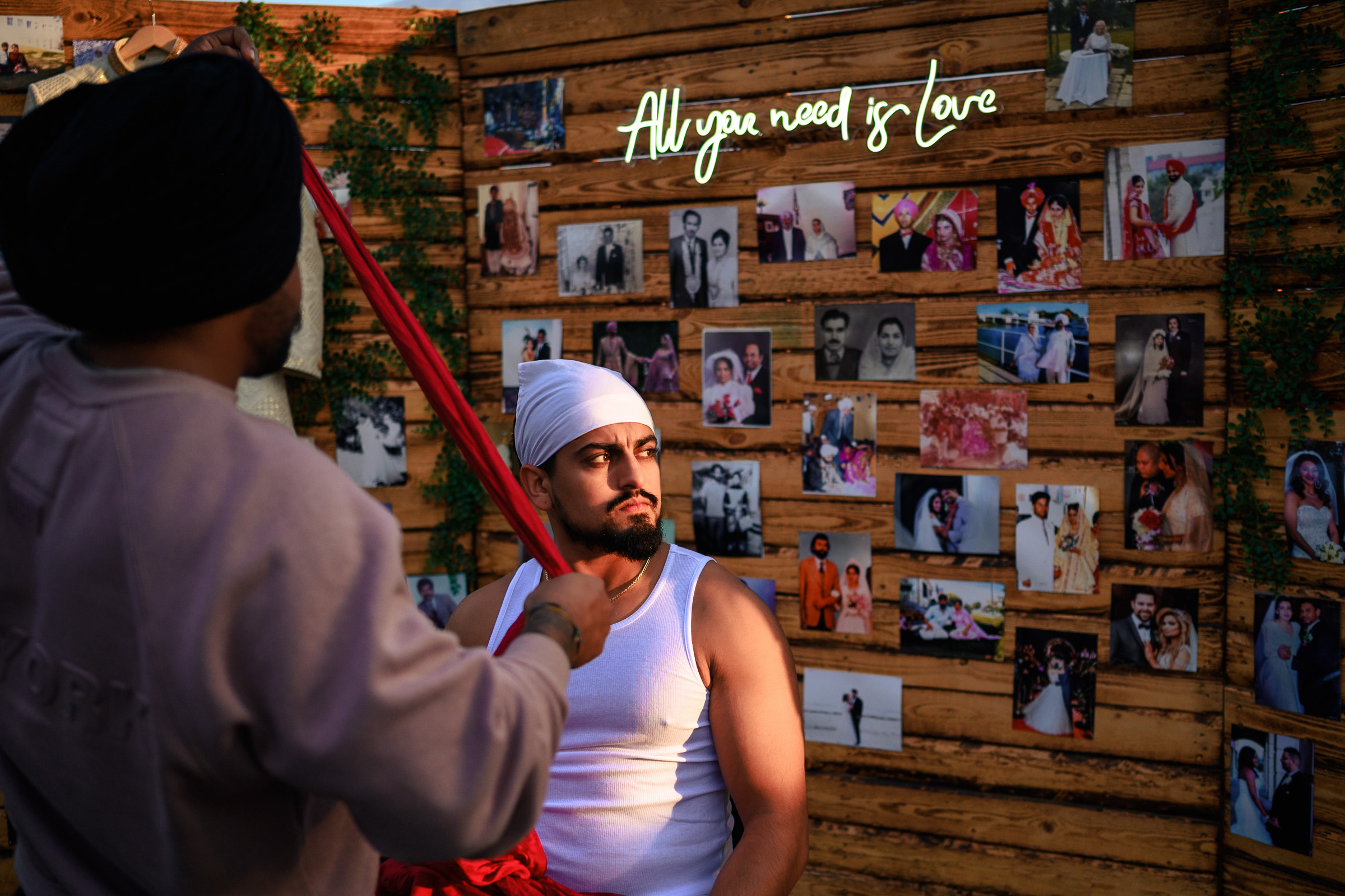 Two men are talking in front of a photo wall with a neon sign reading "All you need is love.