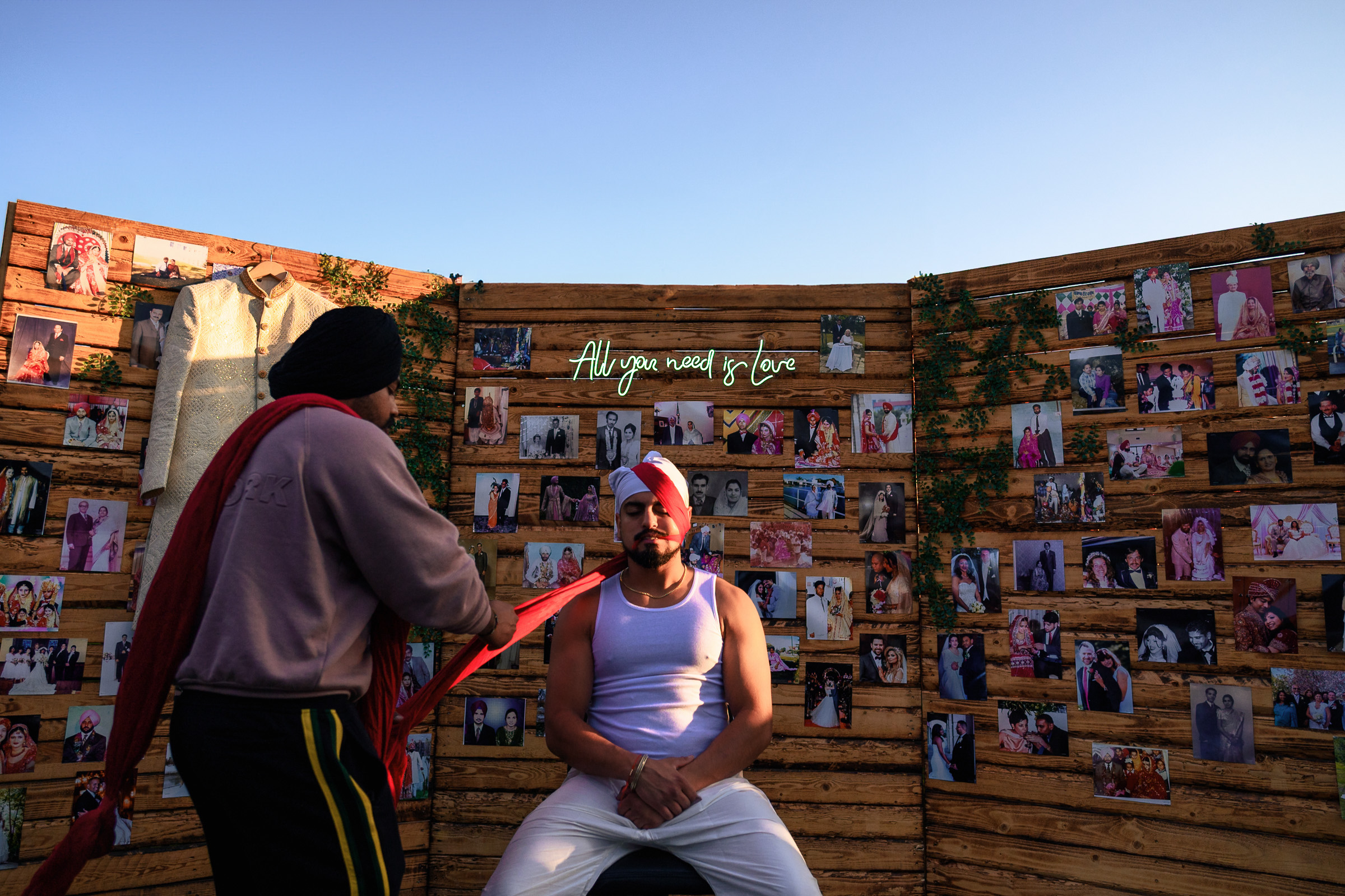 Person having a turban tied, seated against a backdrop of photos and a "All you need is love" sign.
