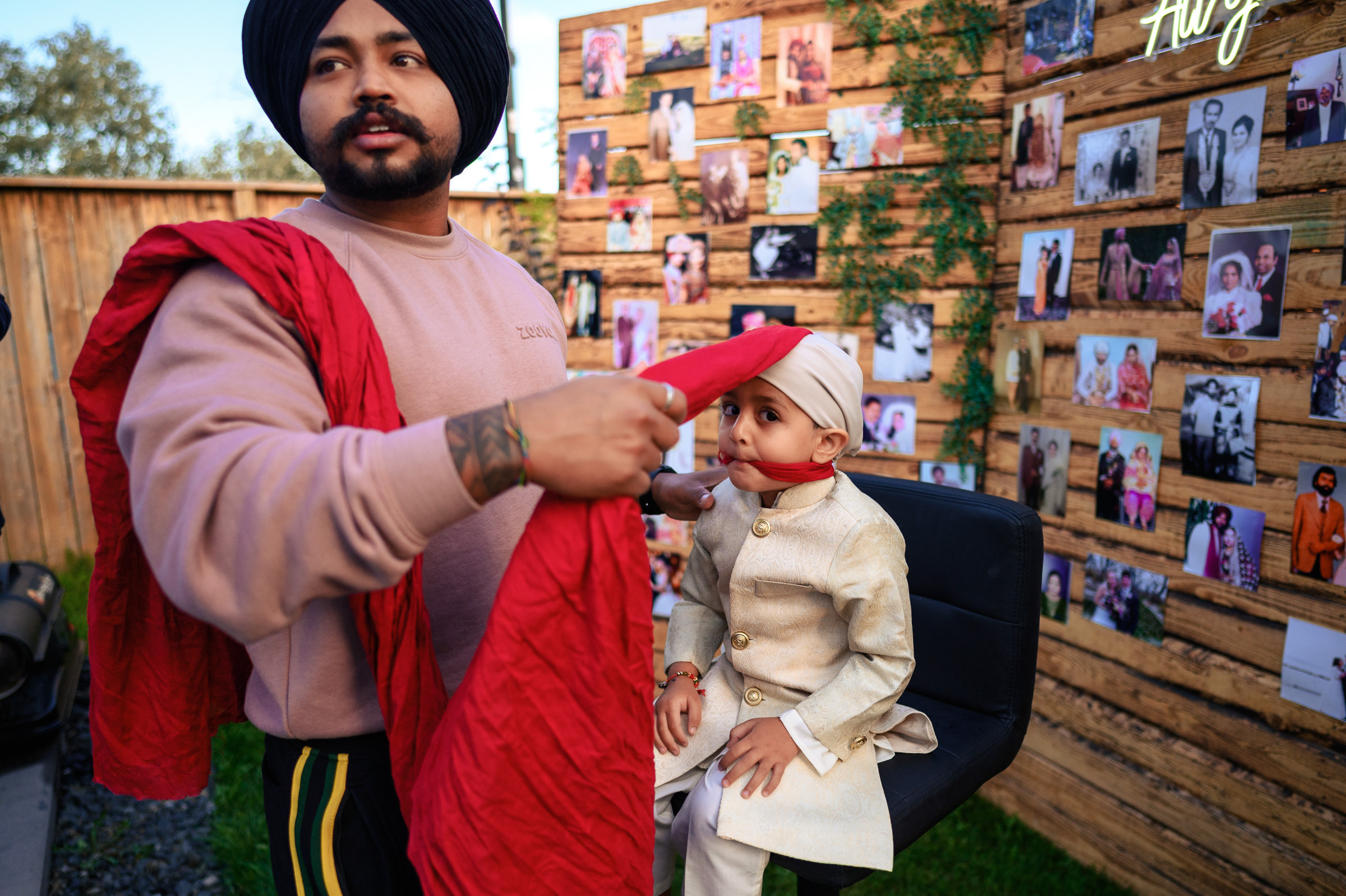 A man ties a turban on a child seated in front of a photo-covered wall.