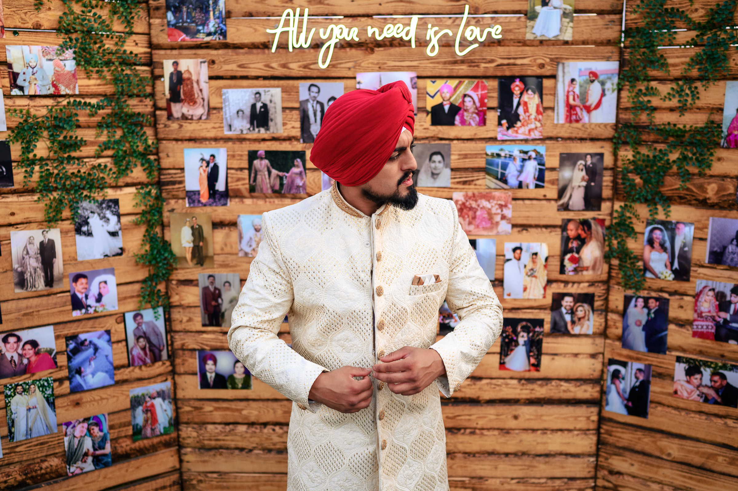 Man in red turban and gold sherwani stands before photos, with "All you need is love" sign above.
