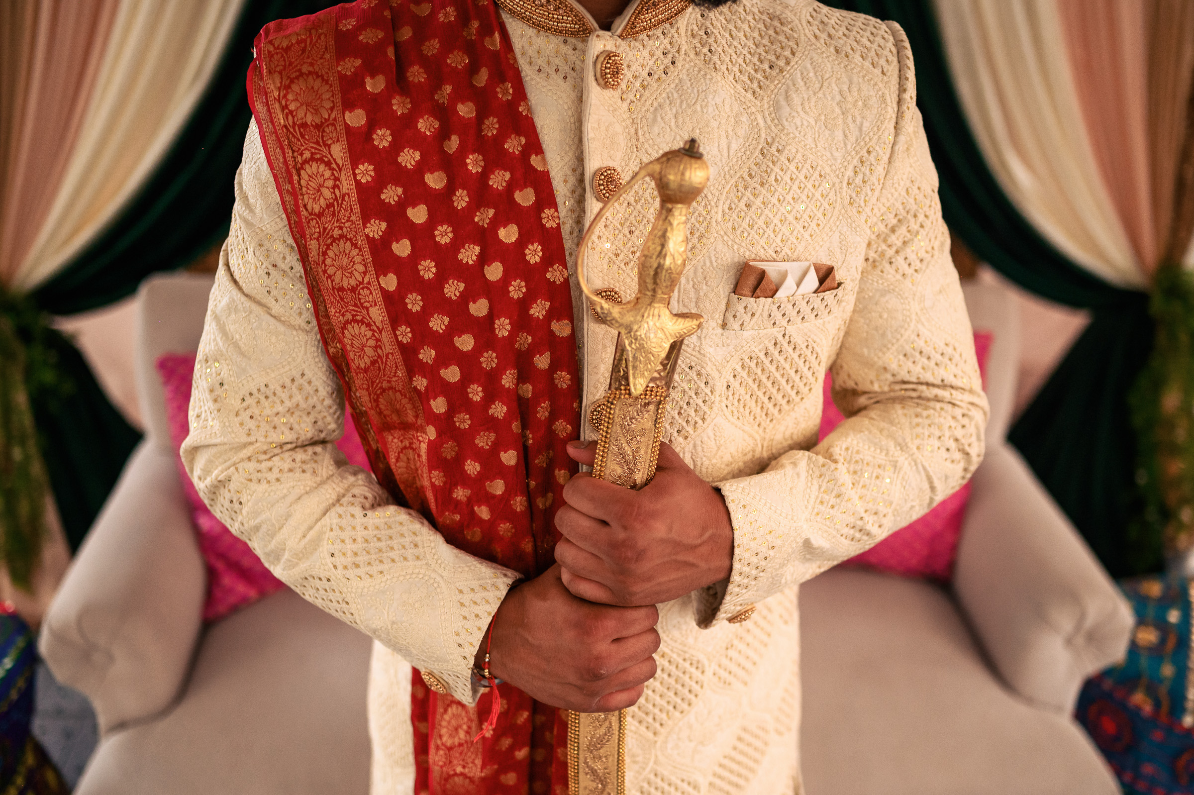 Man in a cream sherwani with a red stole, holding a ceremonial sword.