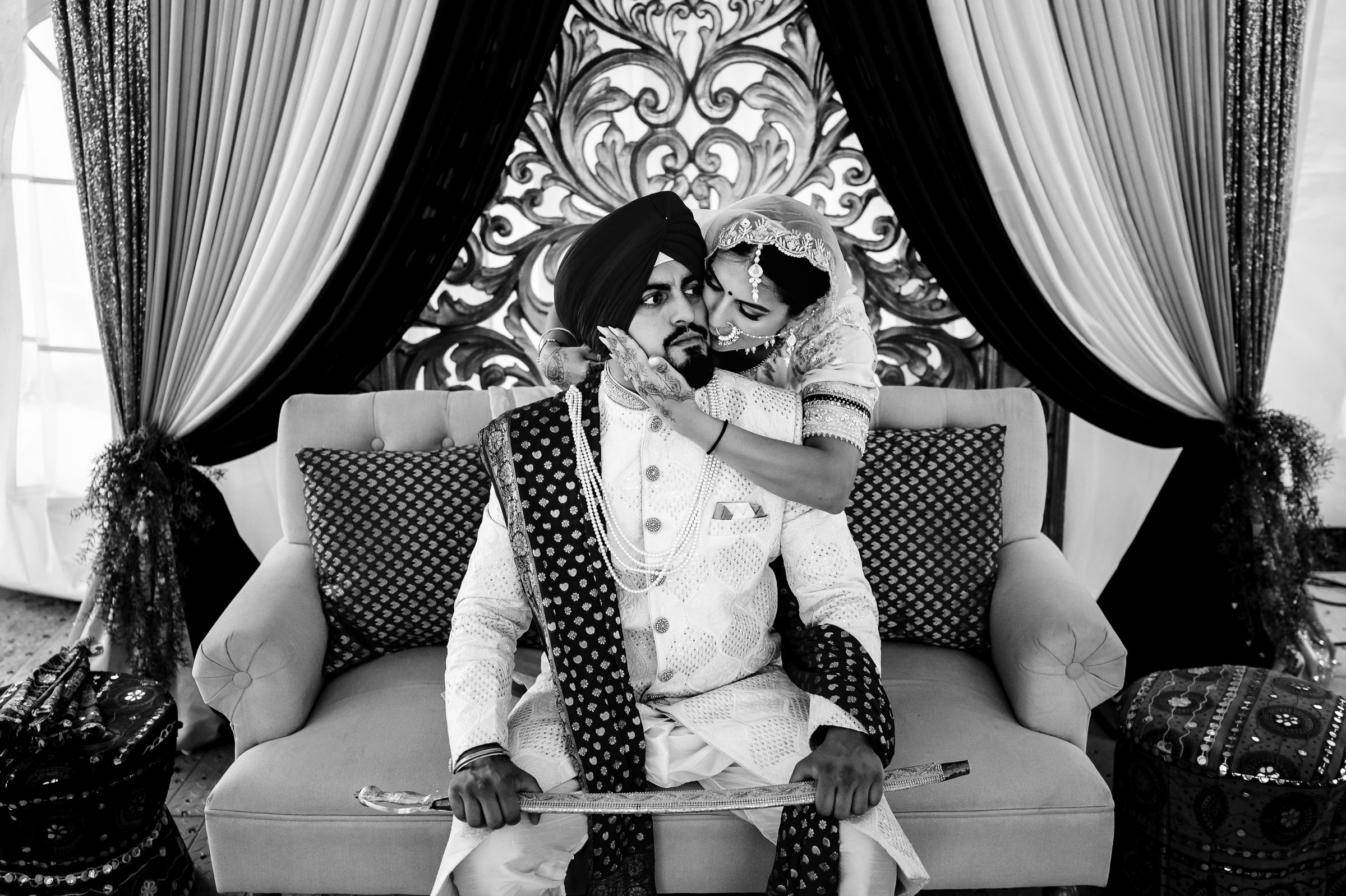 Bride and groom sitting on a couch in ornate traditional attire, embracing.