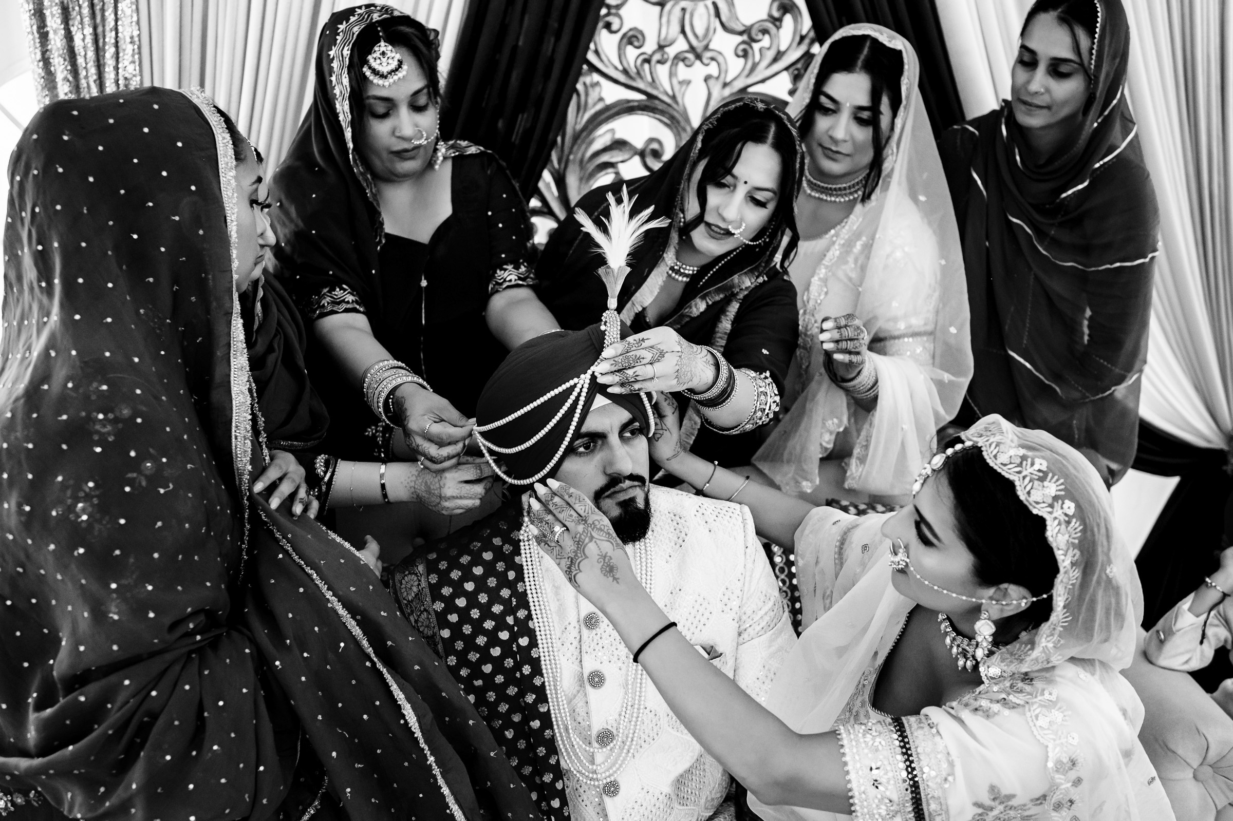 Women assist a groom wearing a turban in a traditional ceremony setting.