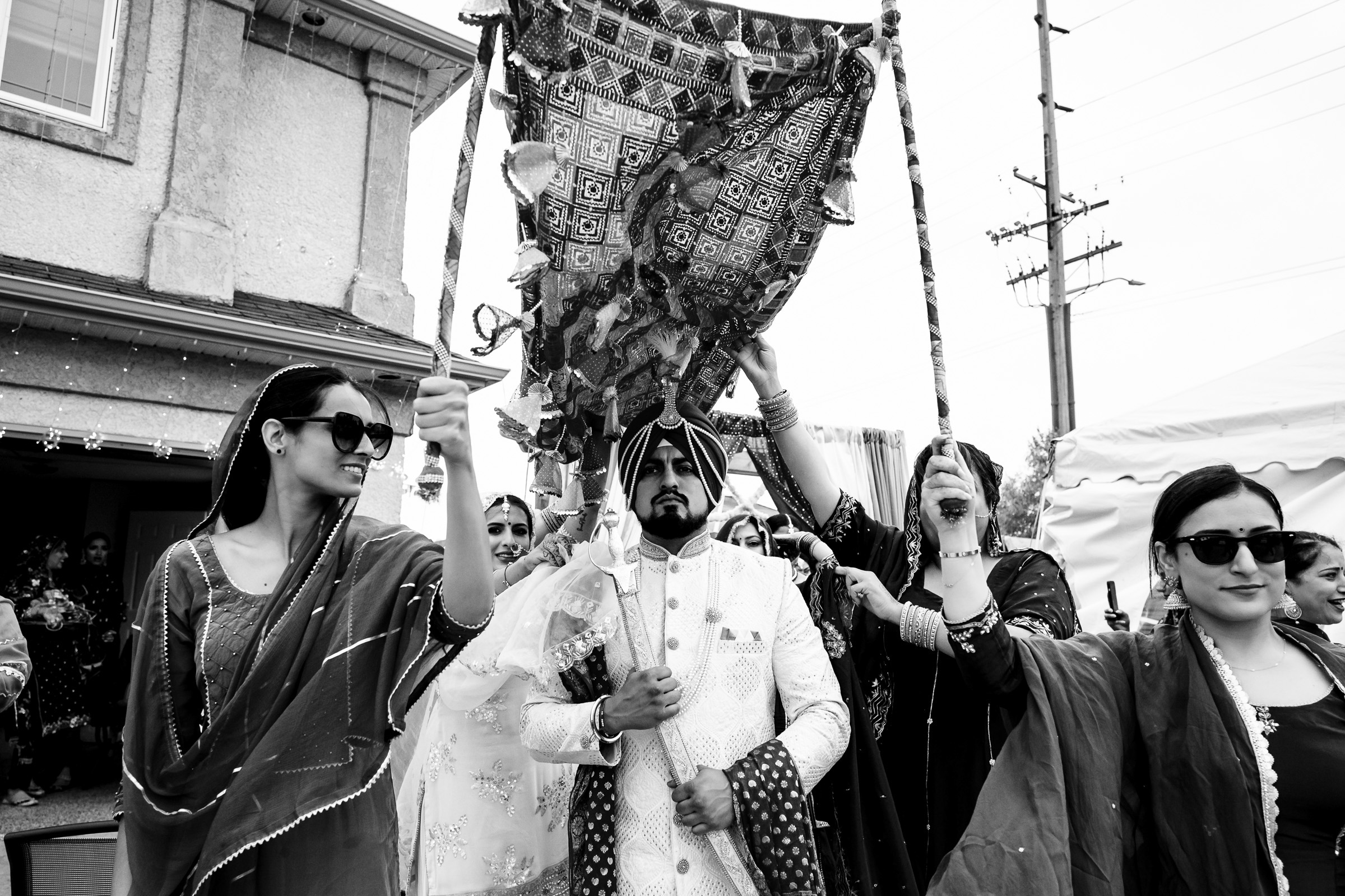 A groom under a traditional canopy, with women holding it up, during a procession.