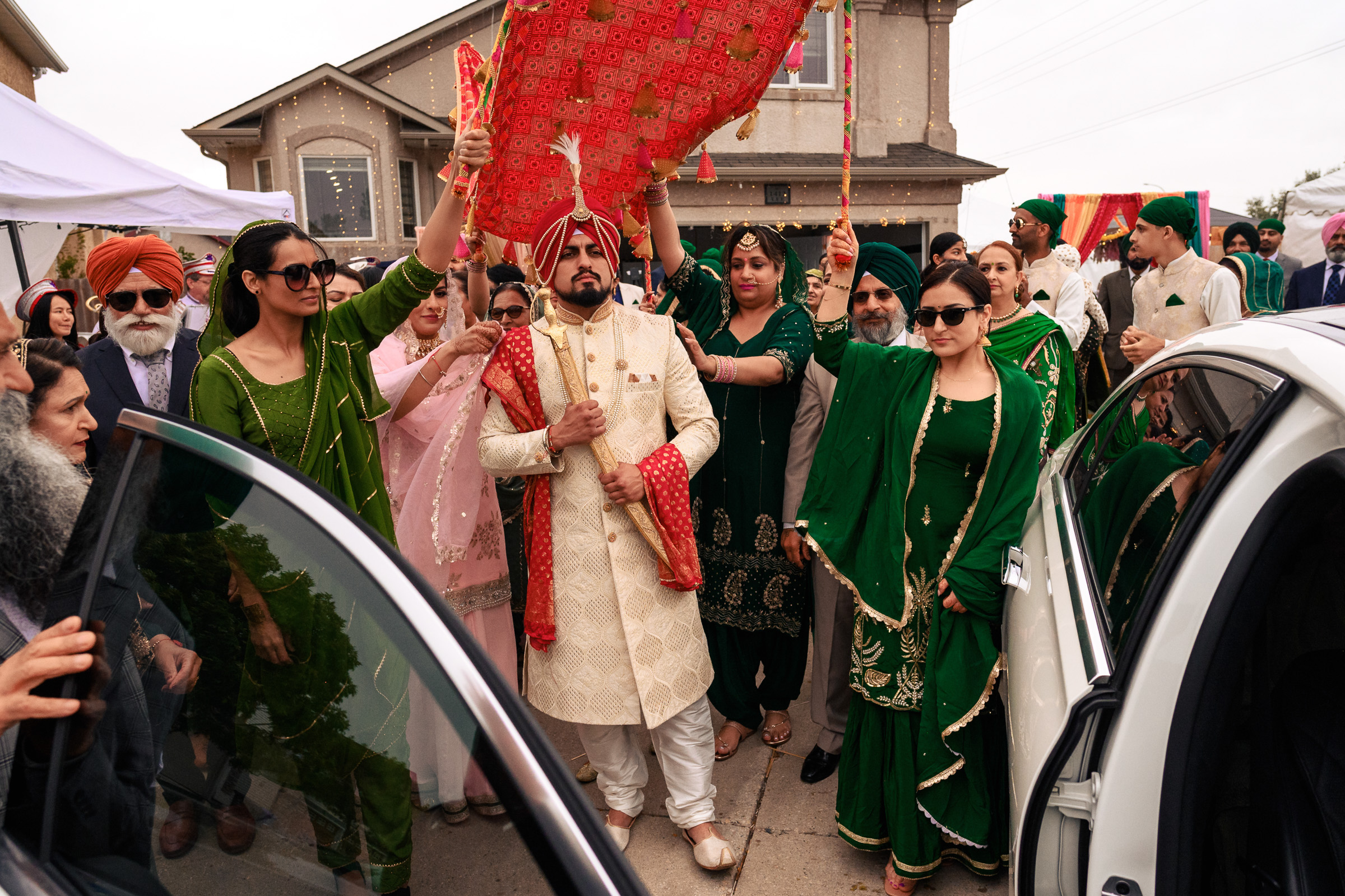 Man in traditional attire, surrounded by people, under a decorative canopy near a car.