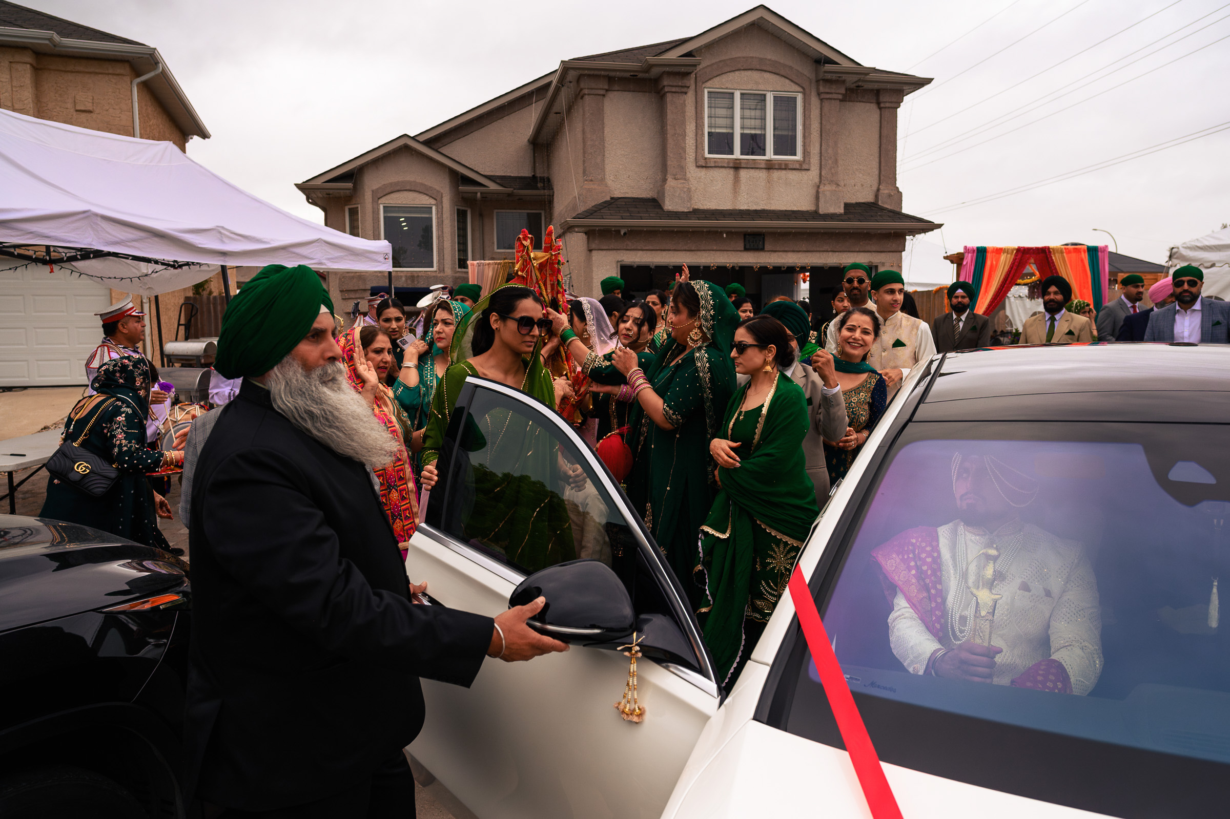 A man opens a car door as a group of people in green attire celebrate outside a house.