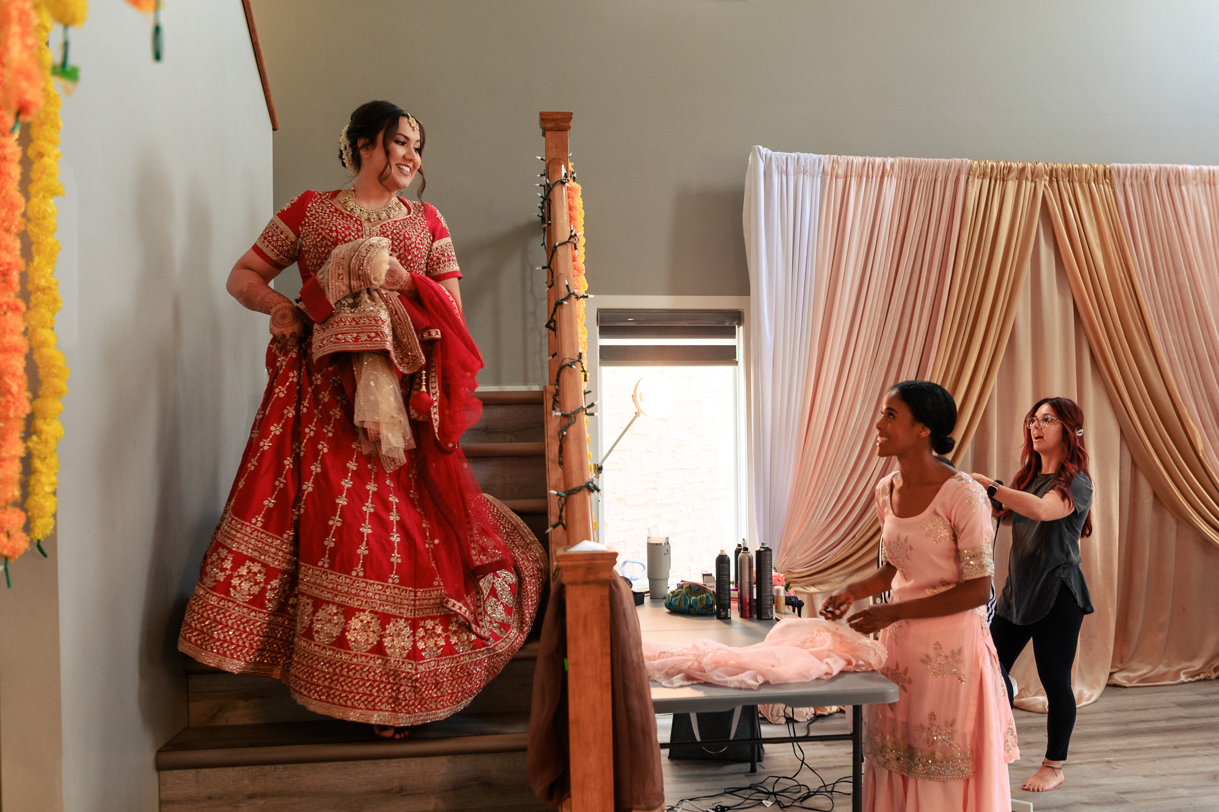Bride in red dress descends stairs; two women assist with garment preparation.
