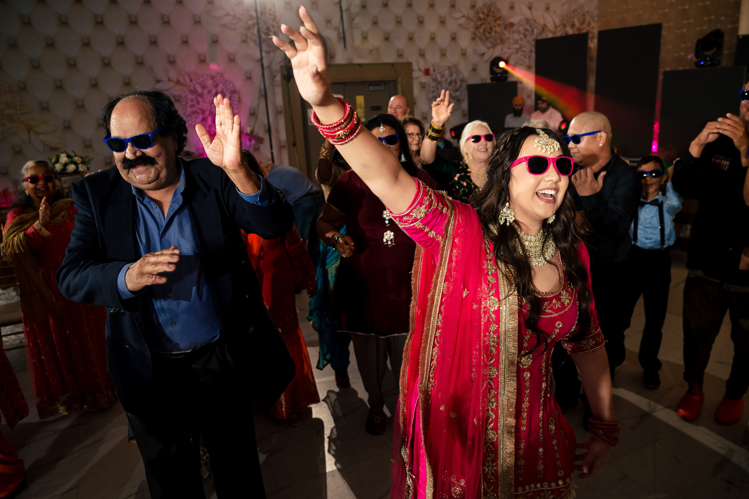 People in colorful attire and sunglasses dancing energetically at a lively indoor event.
