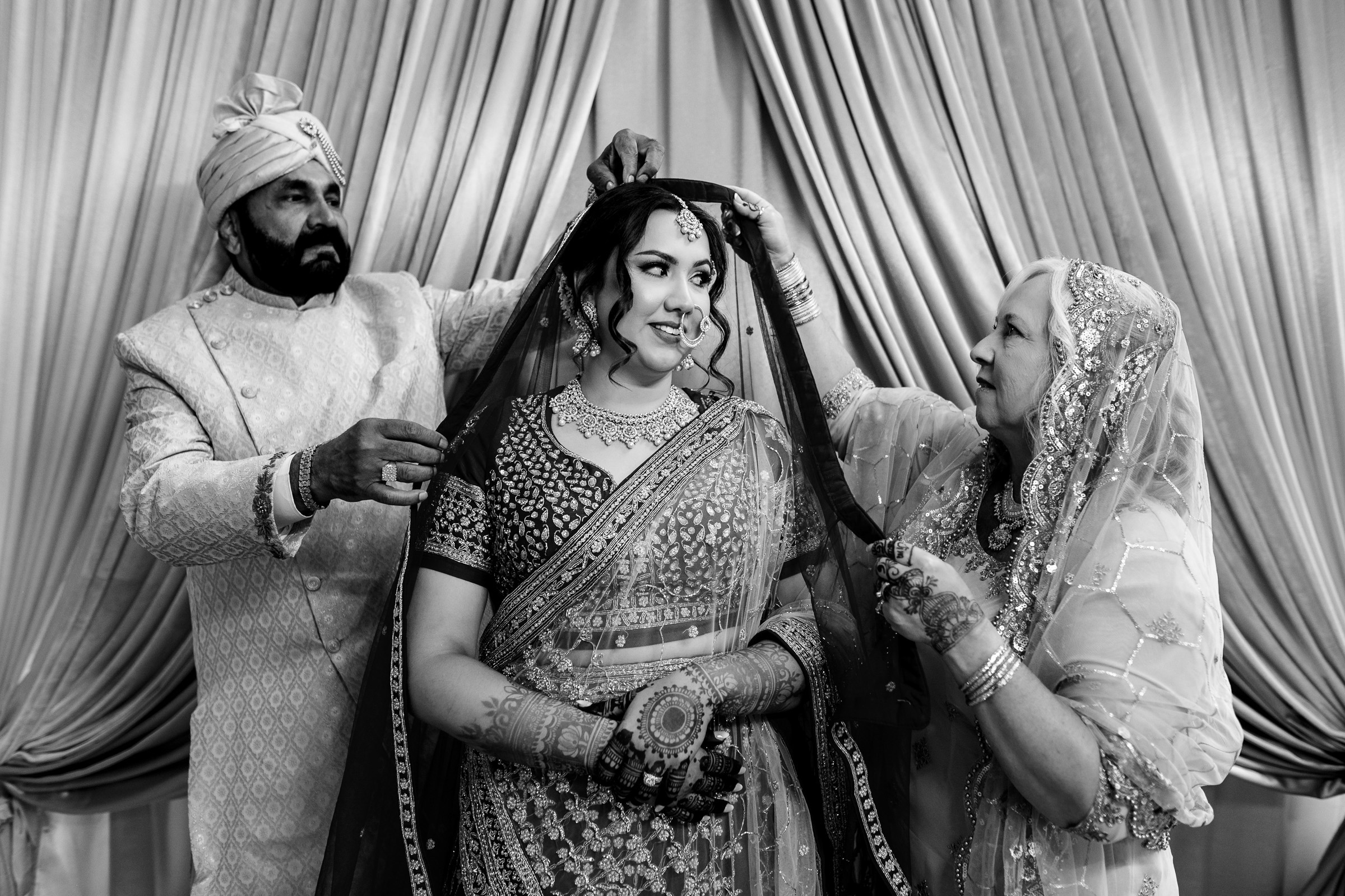Bride in traditional attire with two people adjusting her veil, draped backdrop in background.