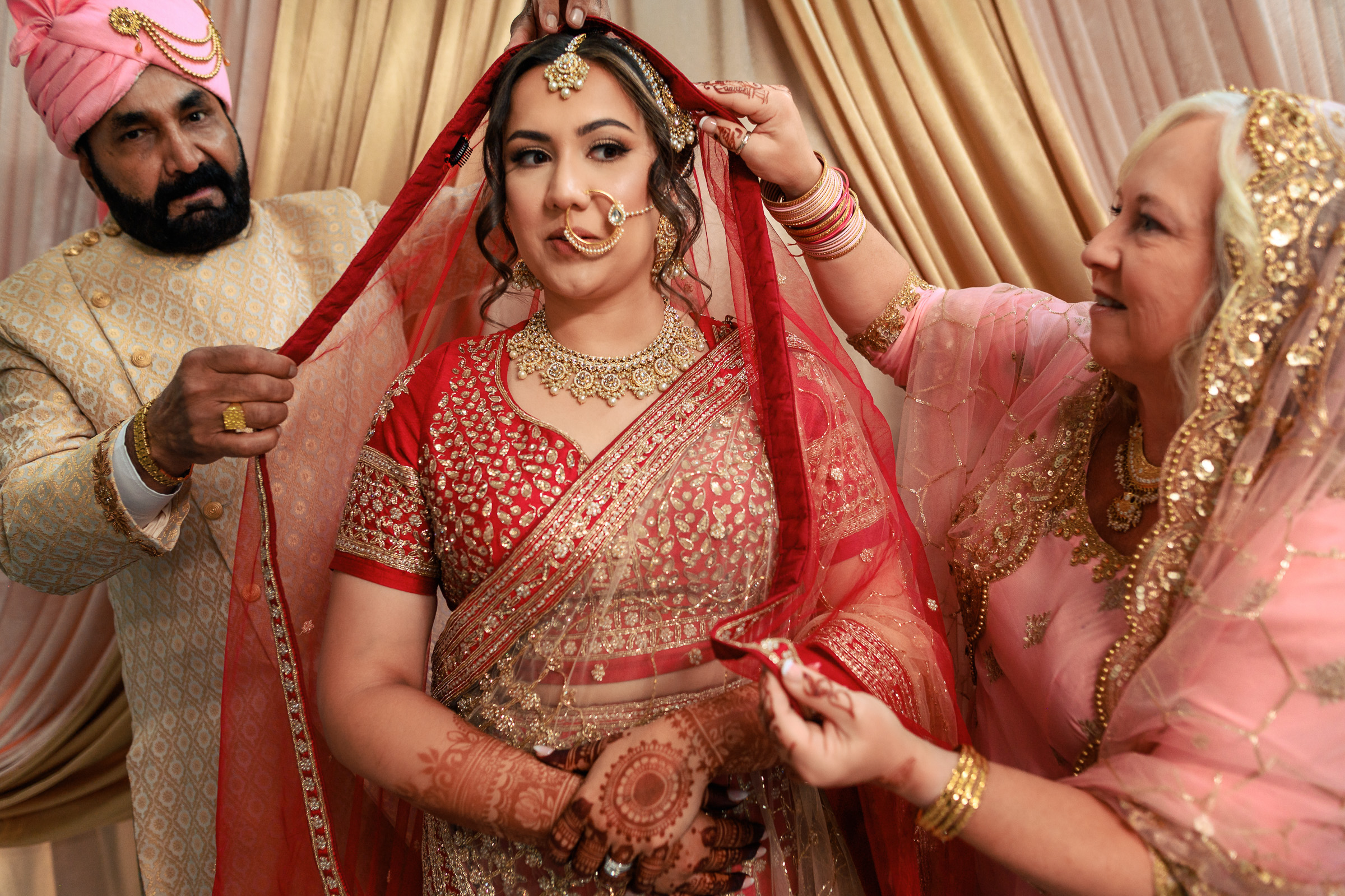 Bride in red traditional attire and jewelry, assisted by two elders fixing her veil.