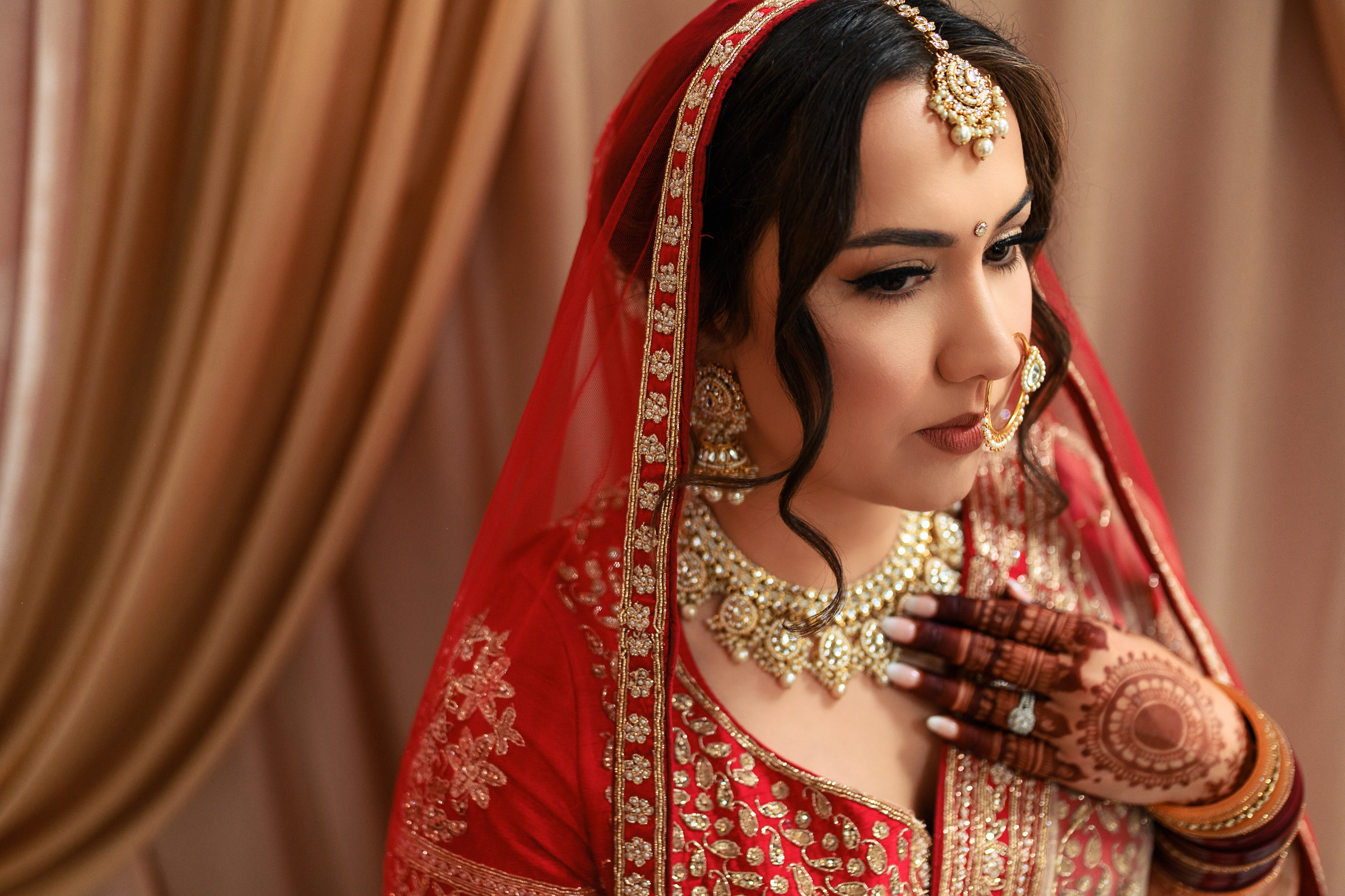Woman in red bridal attire with henna, wearing jewelry, and standing by draped curtains.