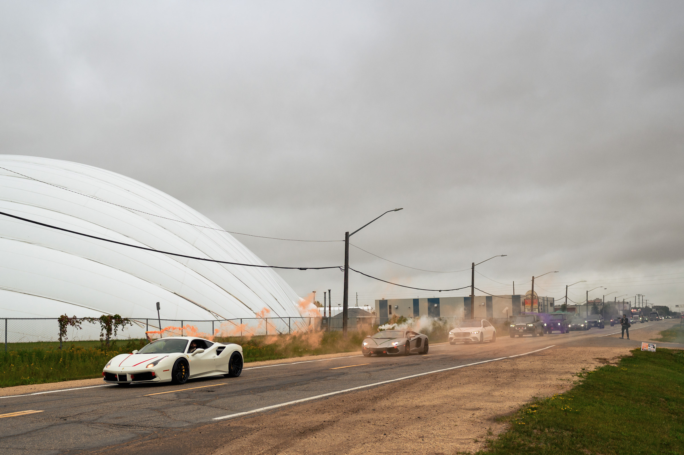 Sports cars driving on a road with colored smoke in the background.