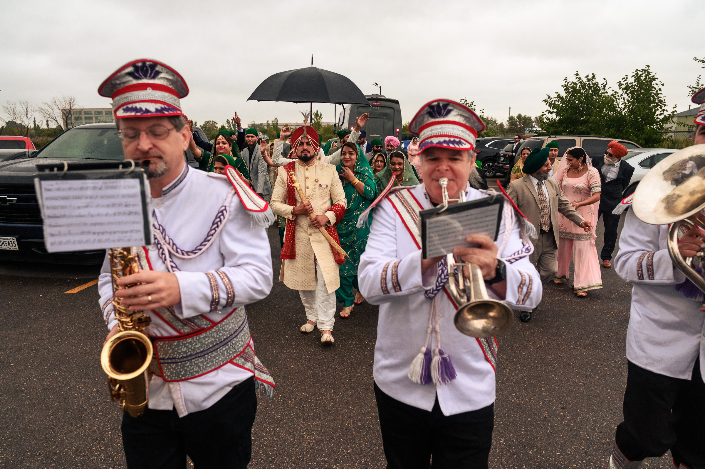 A band leads a joyous wedding procession on a rainy day, with people following under an umbrella.