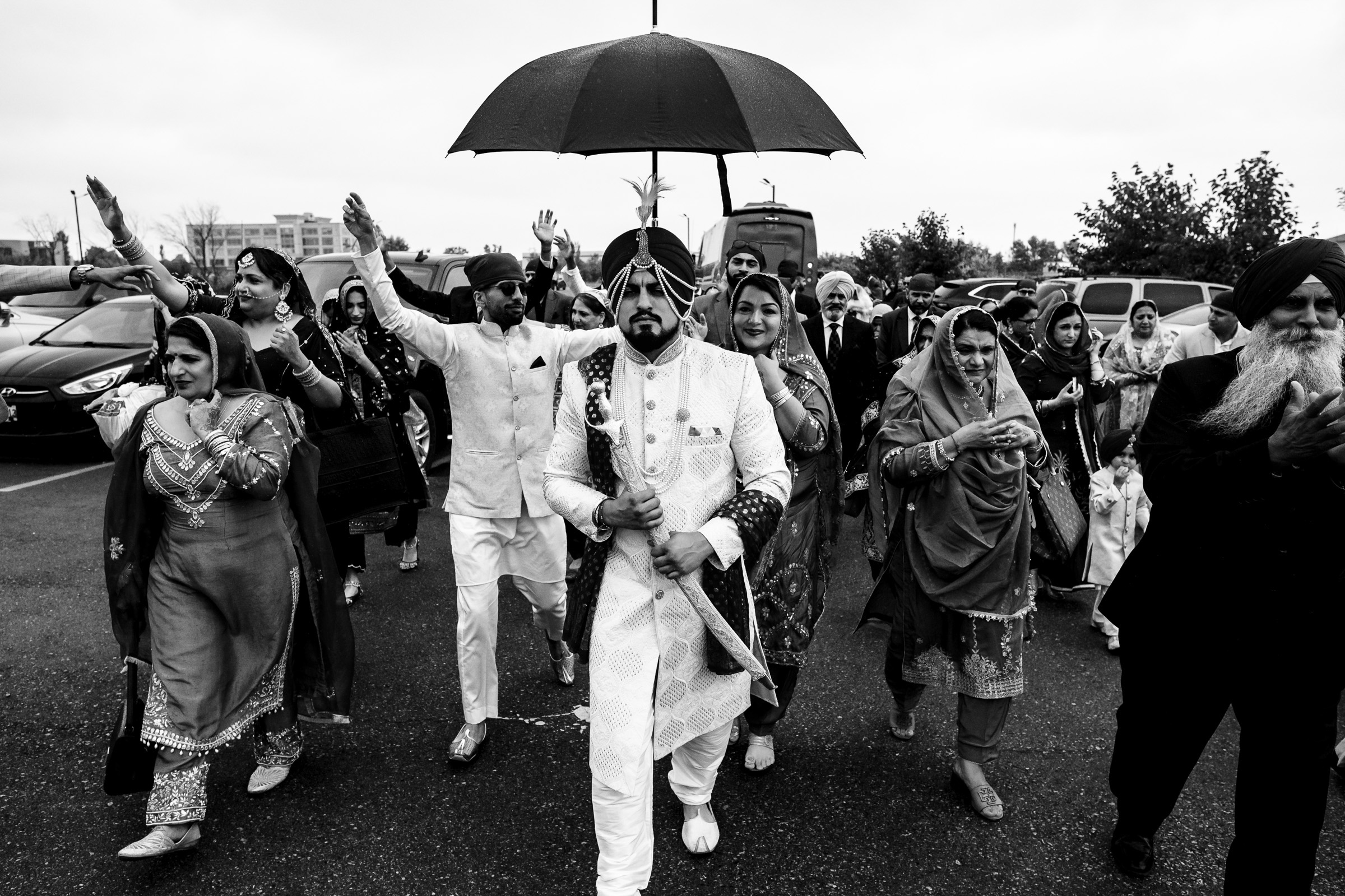 Wedding procession with people in traditional attire, groom holding a ceremonial sword under an umbrella.