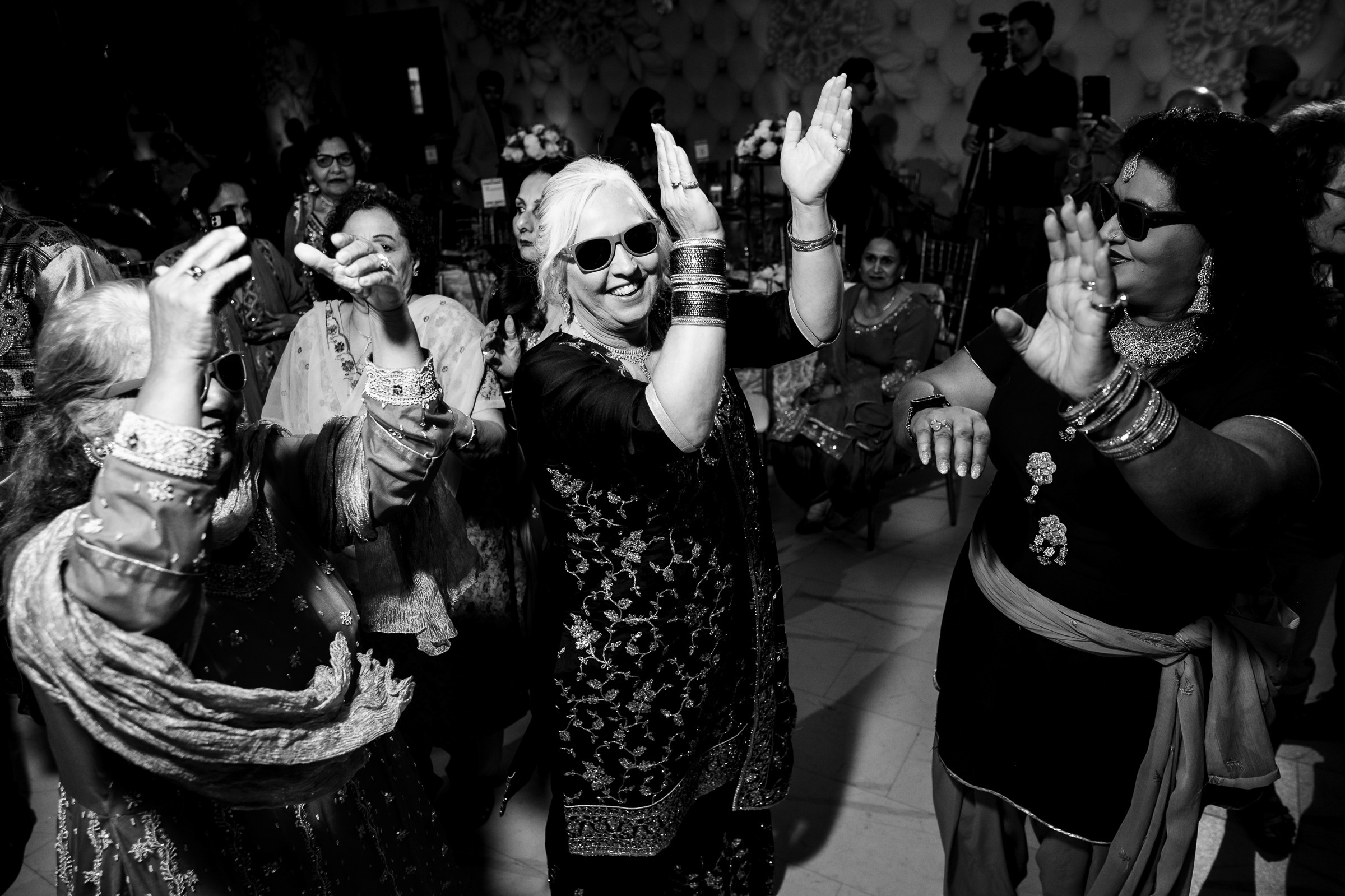 People dancing energetically in traditional attire and sunglasses at a lively indoor event.