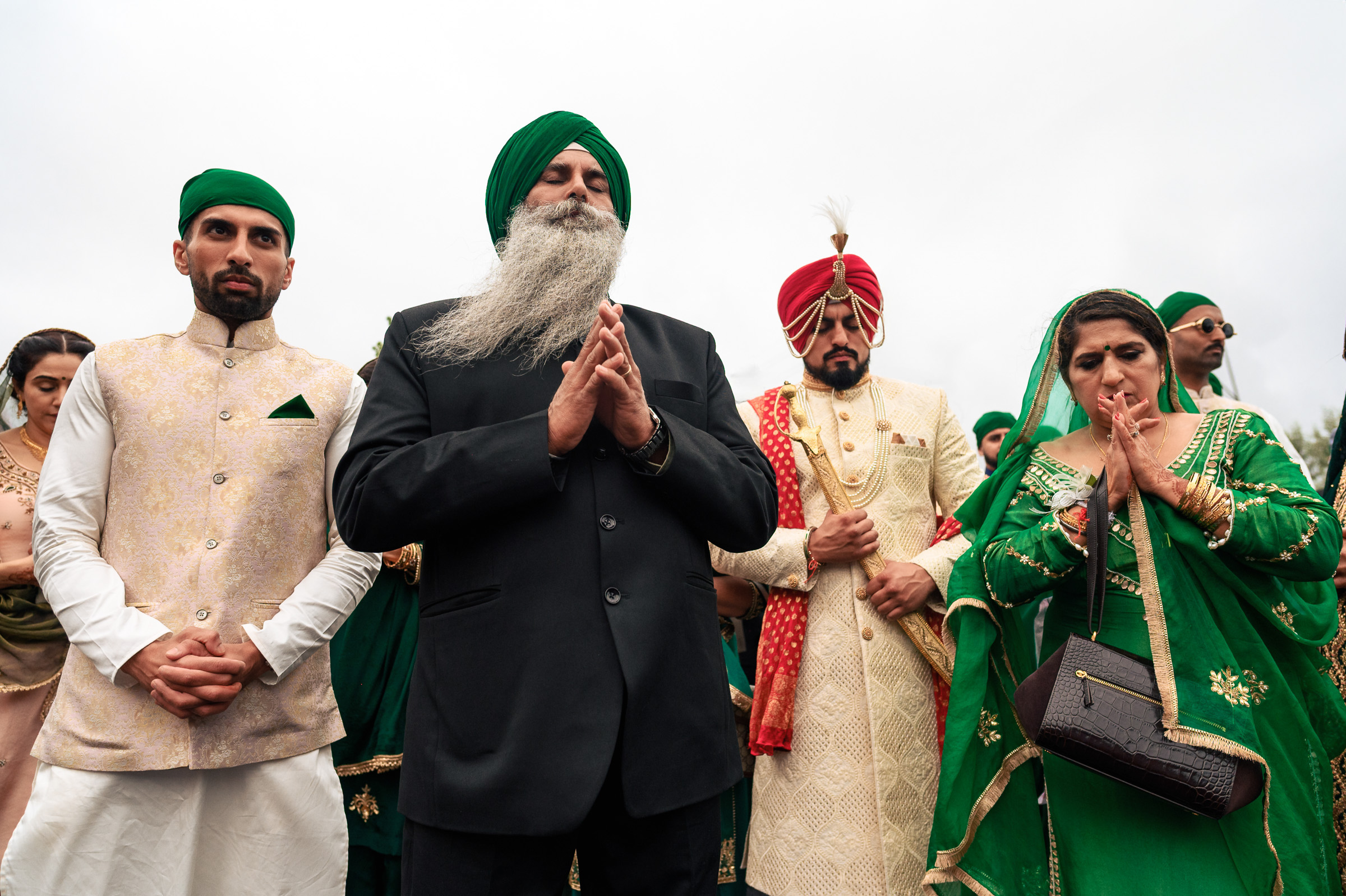 People in traditional attire, standing with hands clasped, outdoors on a cloudy day.