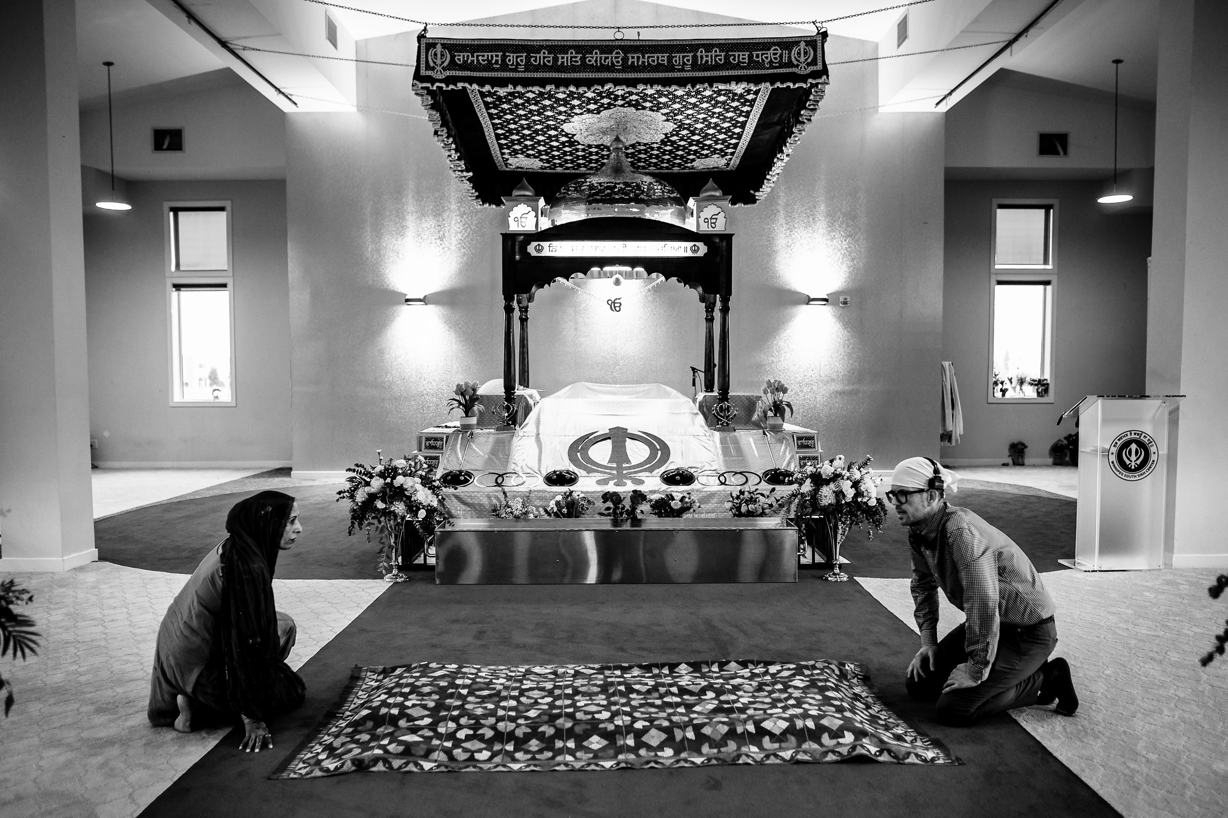 Two people kneeling in a Gurdwara near a religious altar, with floral decor.