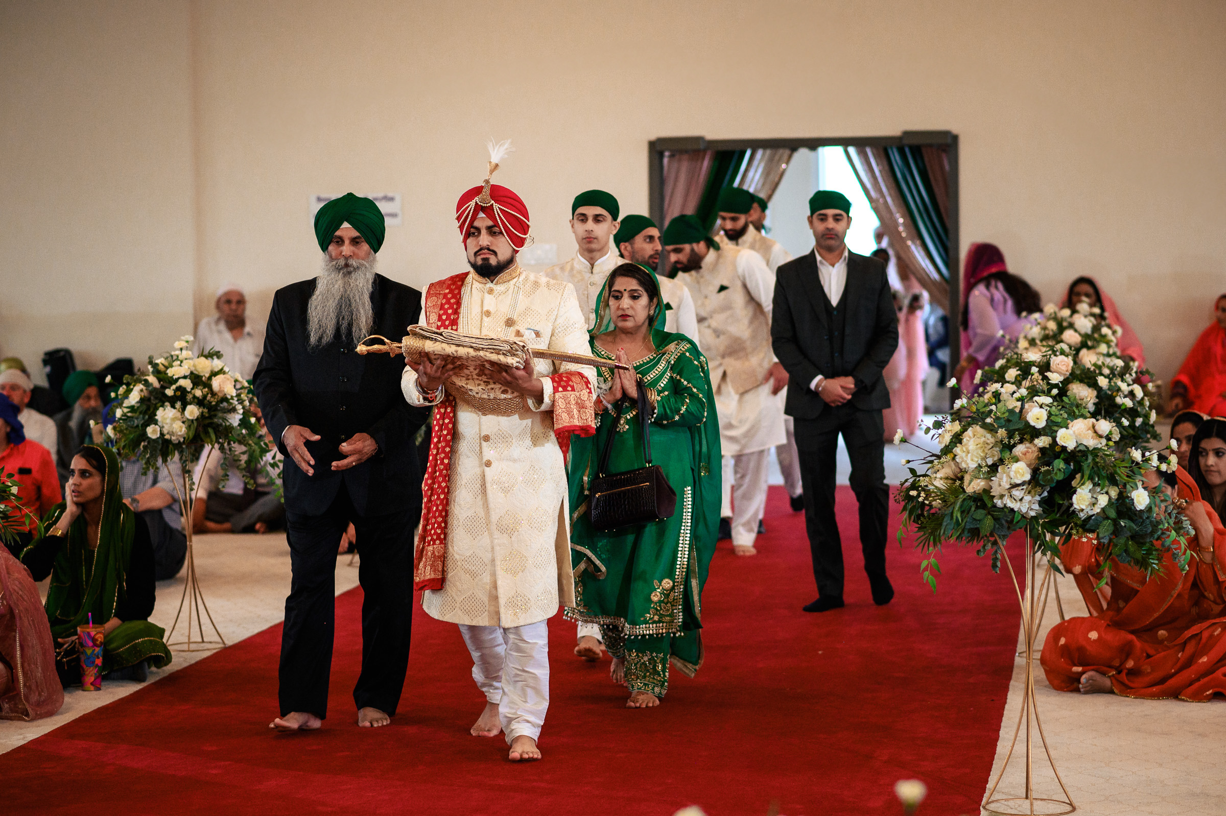 A groom in traditional attire walks down a red carpet, accompanied by family members.