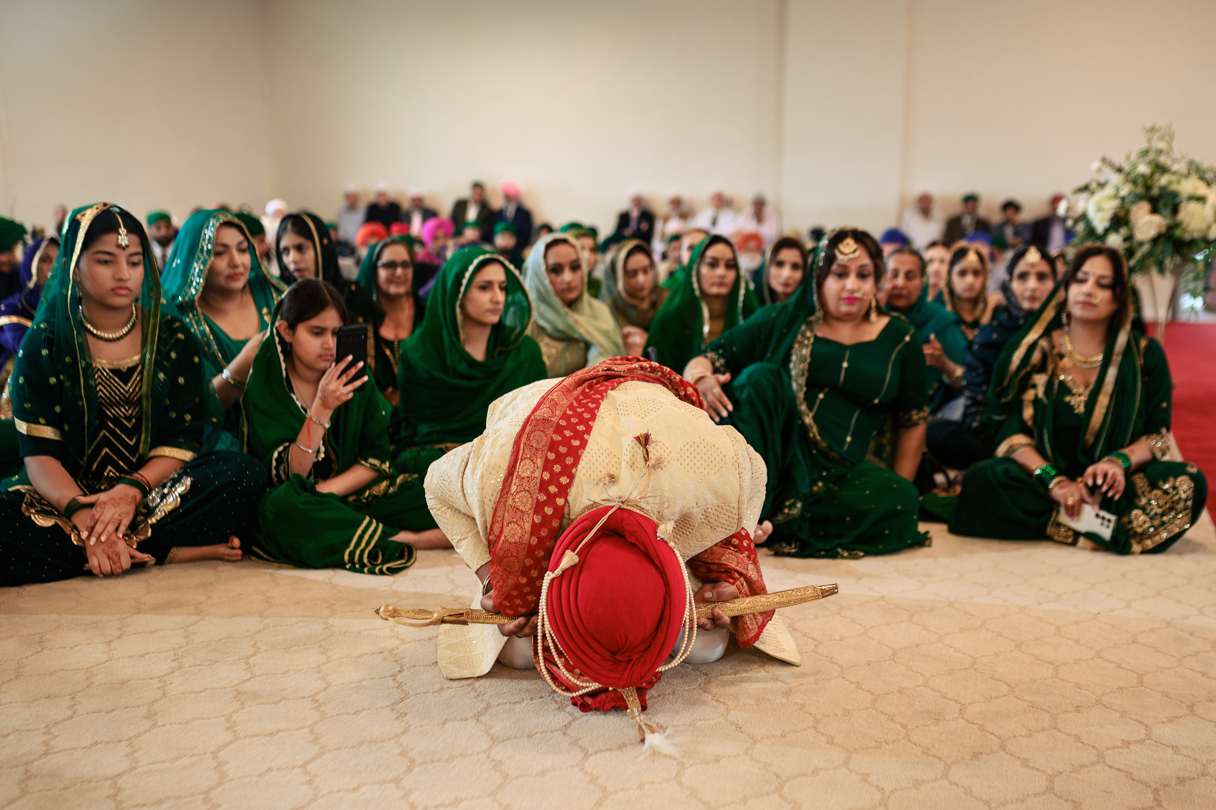 Person in ceremonial attire bows, surrounded by seated people in green clothing.