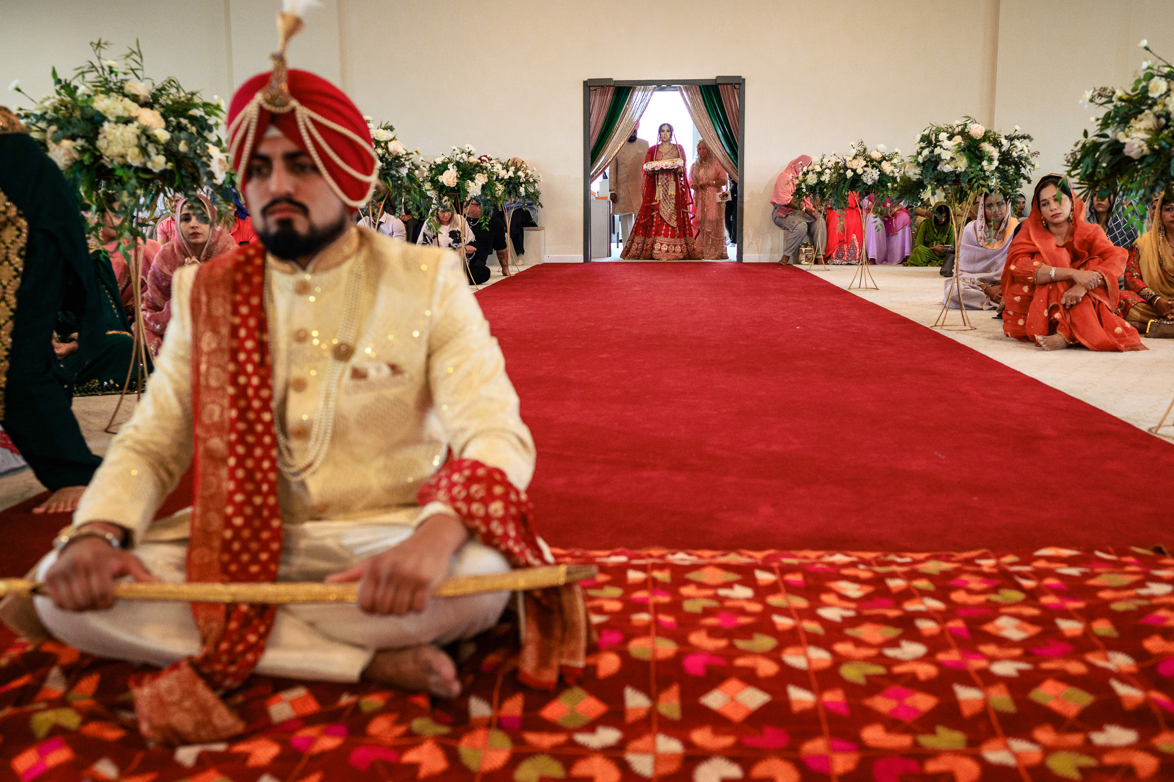 A seated groom in traditional attire; bride enters from the back in a ceremonial setting.