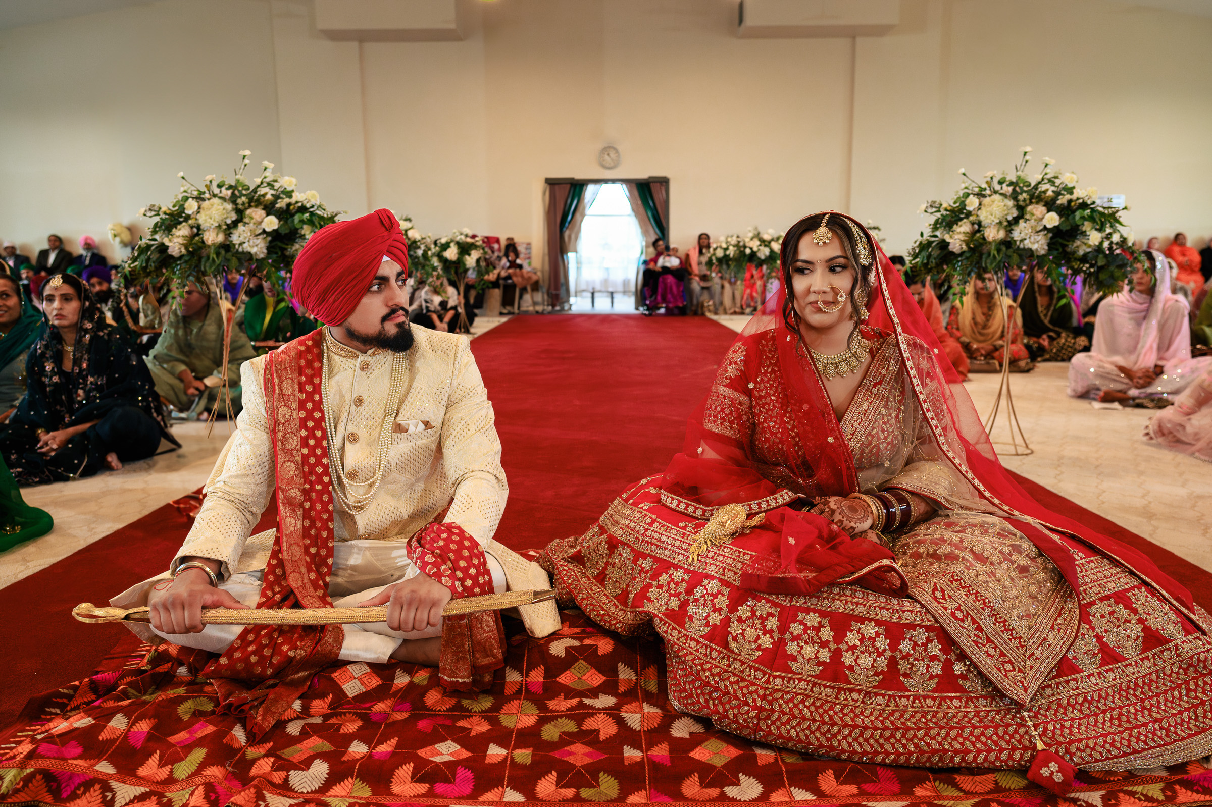A couple in traditional Indian attire sit in a decorated wedding hall.