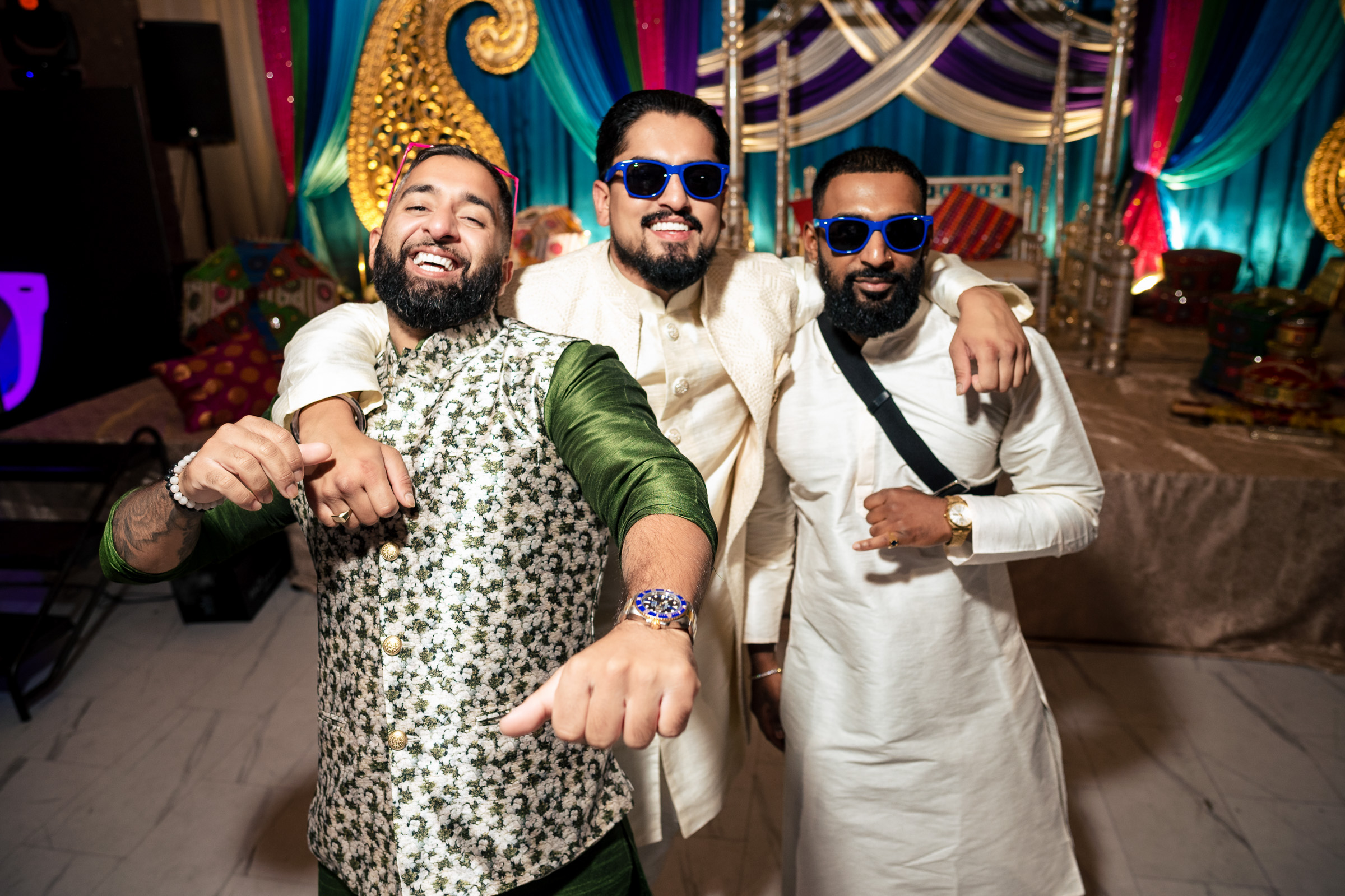 Three men in festive attire pose together, with one wearing sunglasses, against a colorful backdrop.