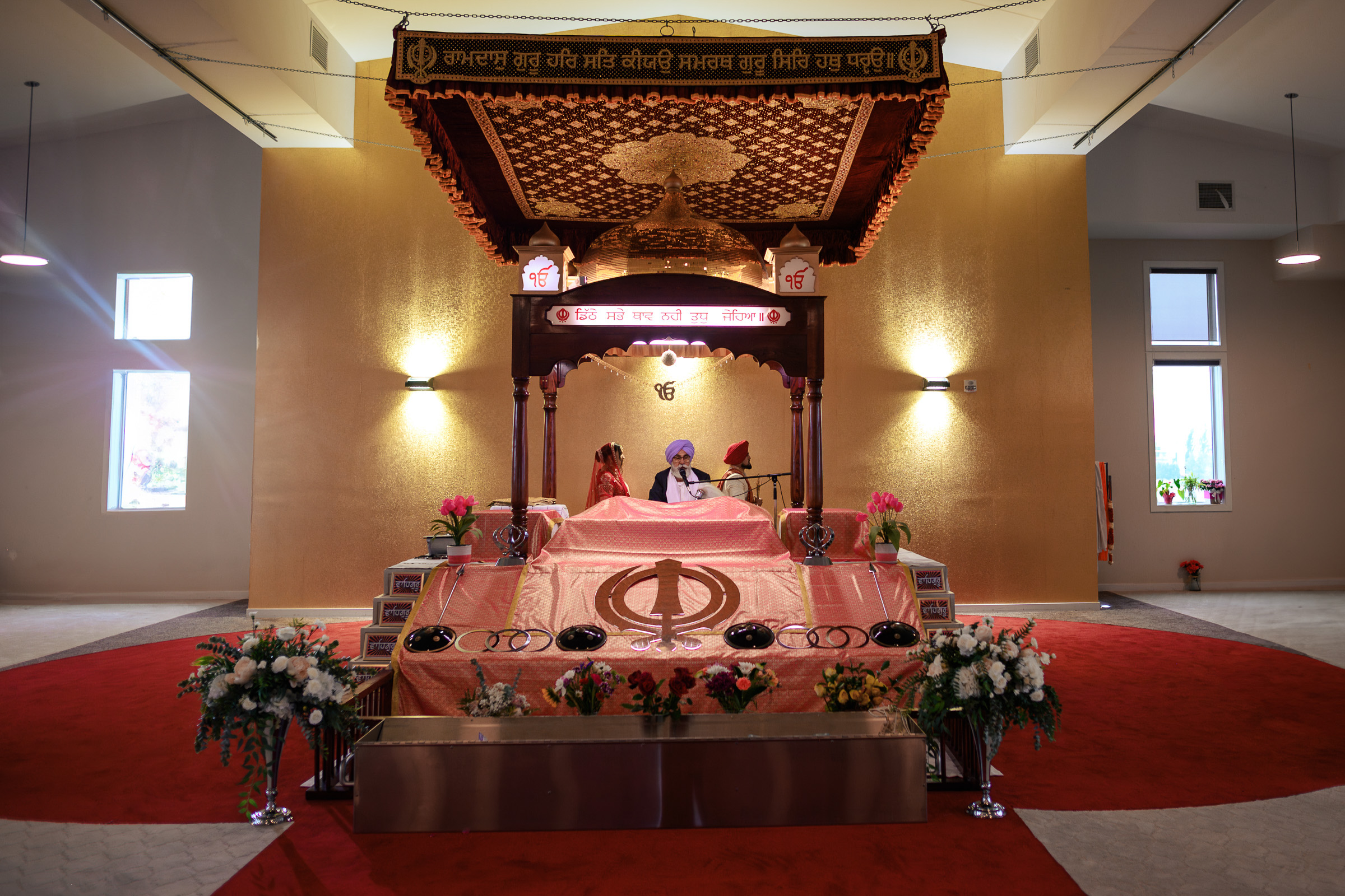 Interior of a gurdwara with two people seated near the Guru Granth Sahib on a decorated platform.