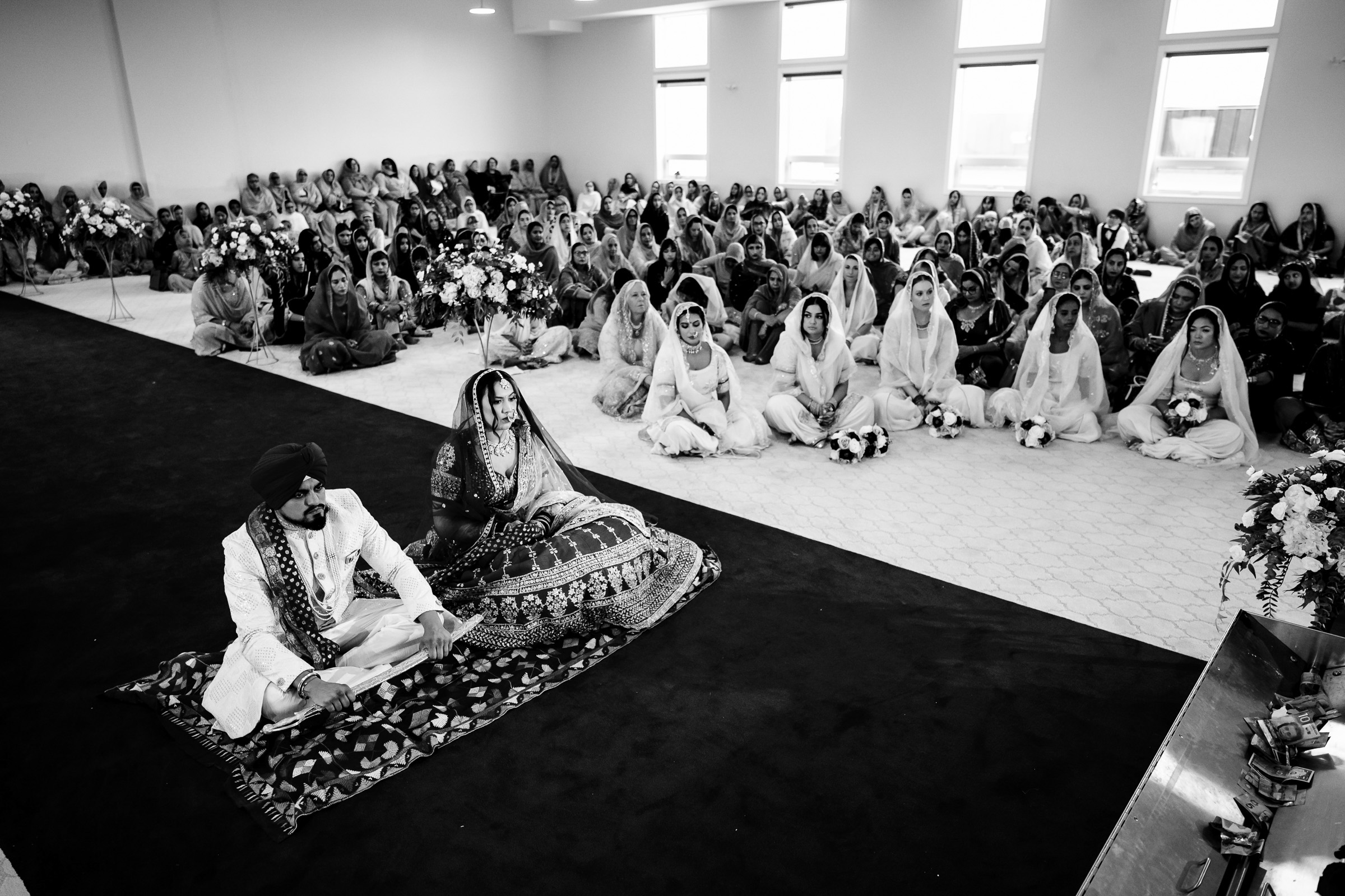 A couple at a traditional ceremony with a seated audience in a decorated hall.