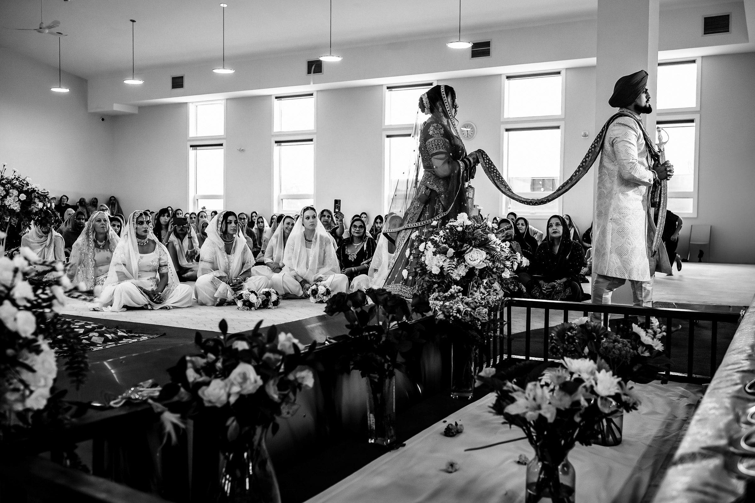 A couple in traditional attire during a wedding ceremony, surrounded by seated guests.