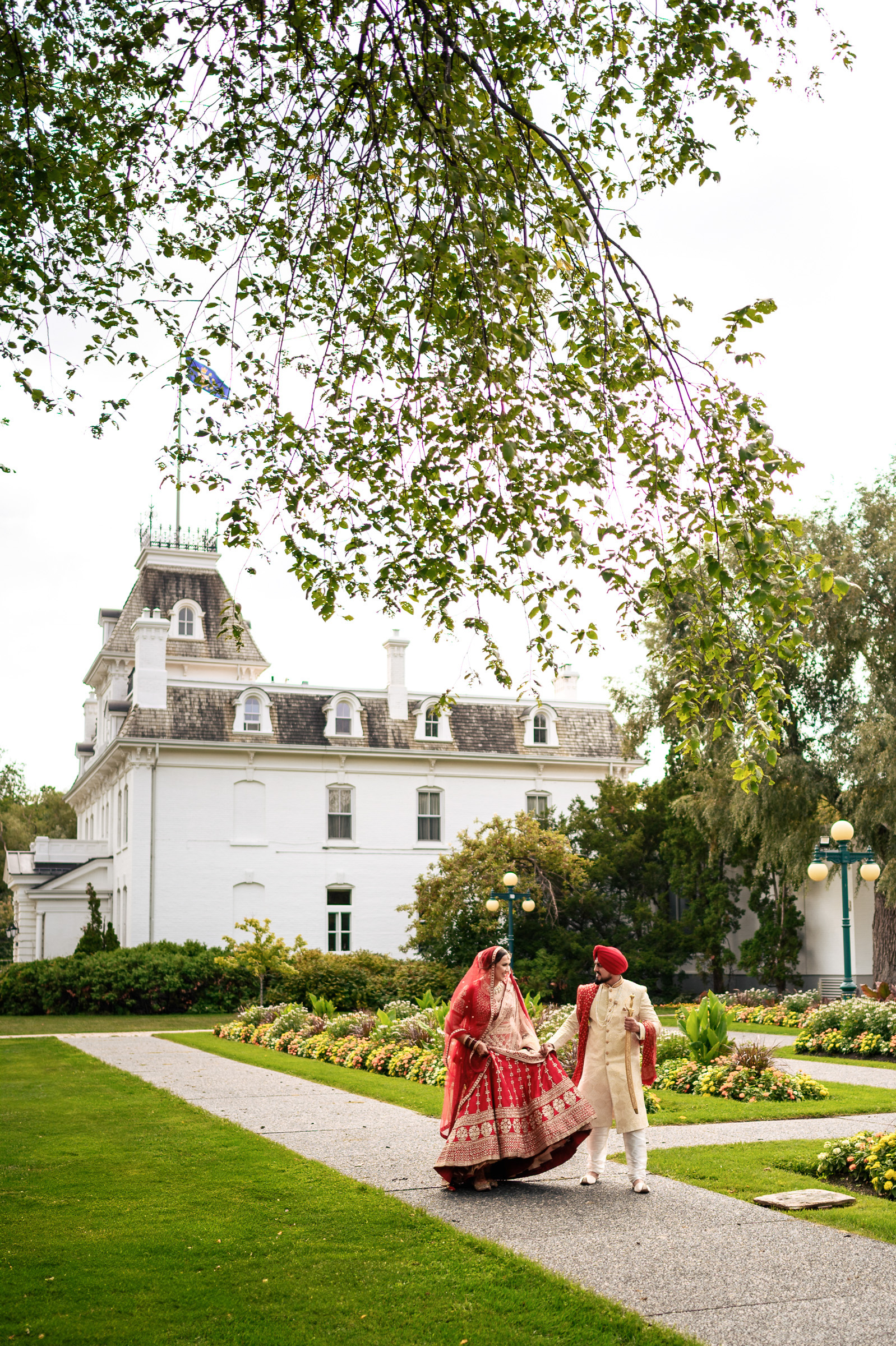 A couple in traditional attire walks on a garden path in front of a large white building.