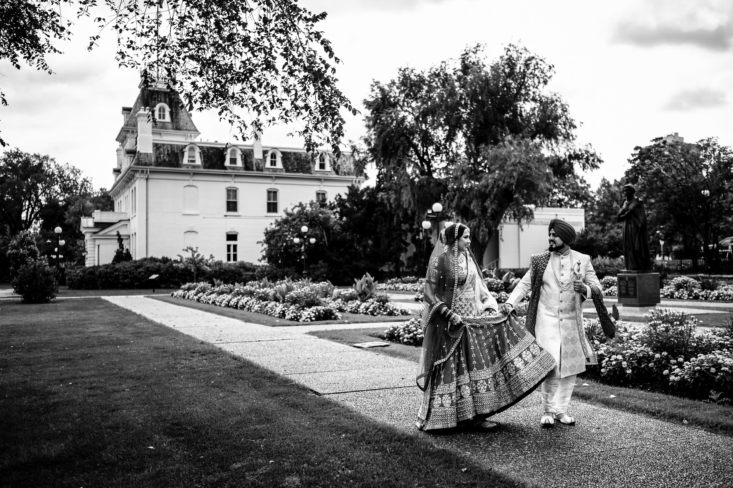 Bride and groom in traditional attire walking on a path near a historic building.