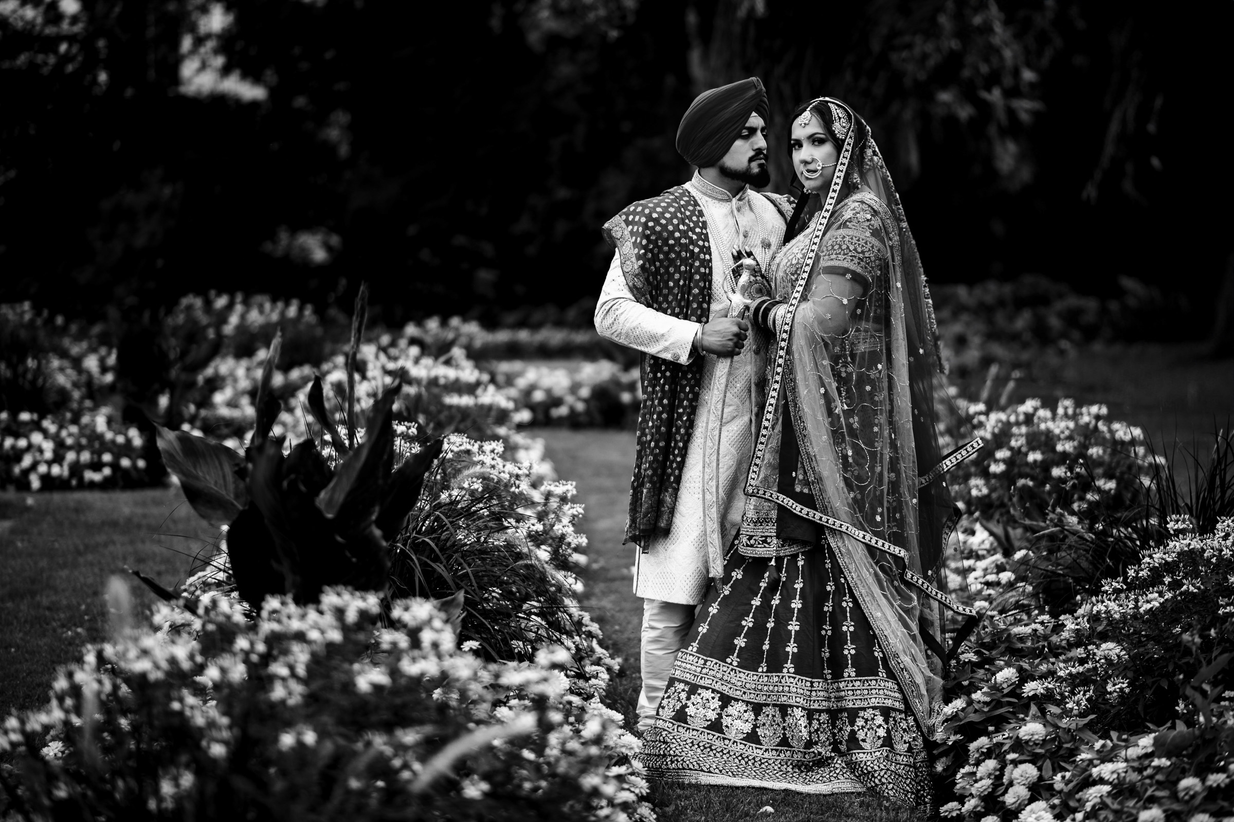 Couple in traditional attire poses in a garden surrounded by flowers.