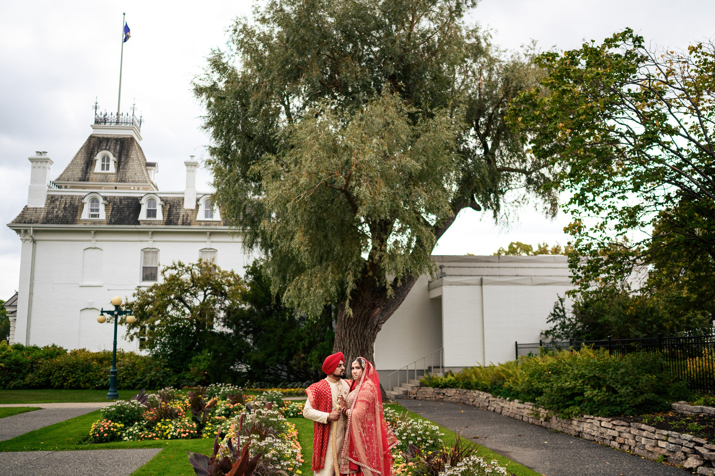 Bride and groom in traditional attire standing in front of a large tree and historic building.