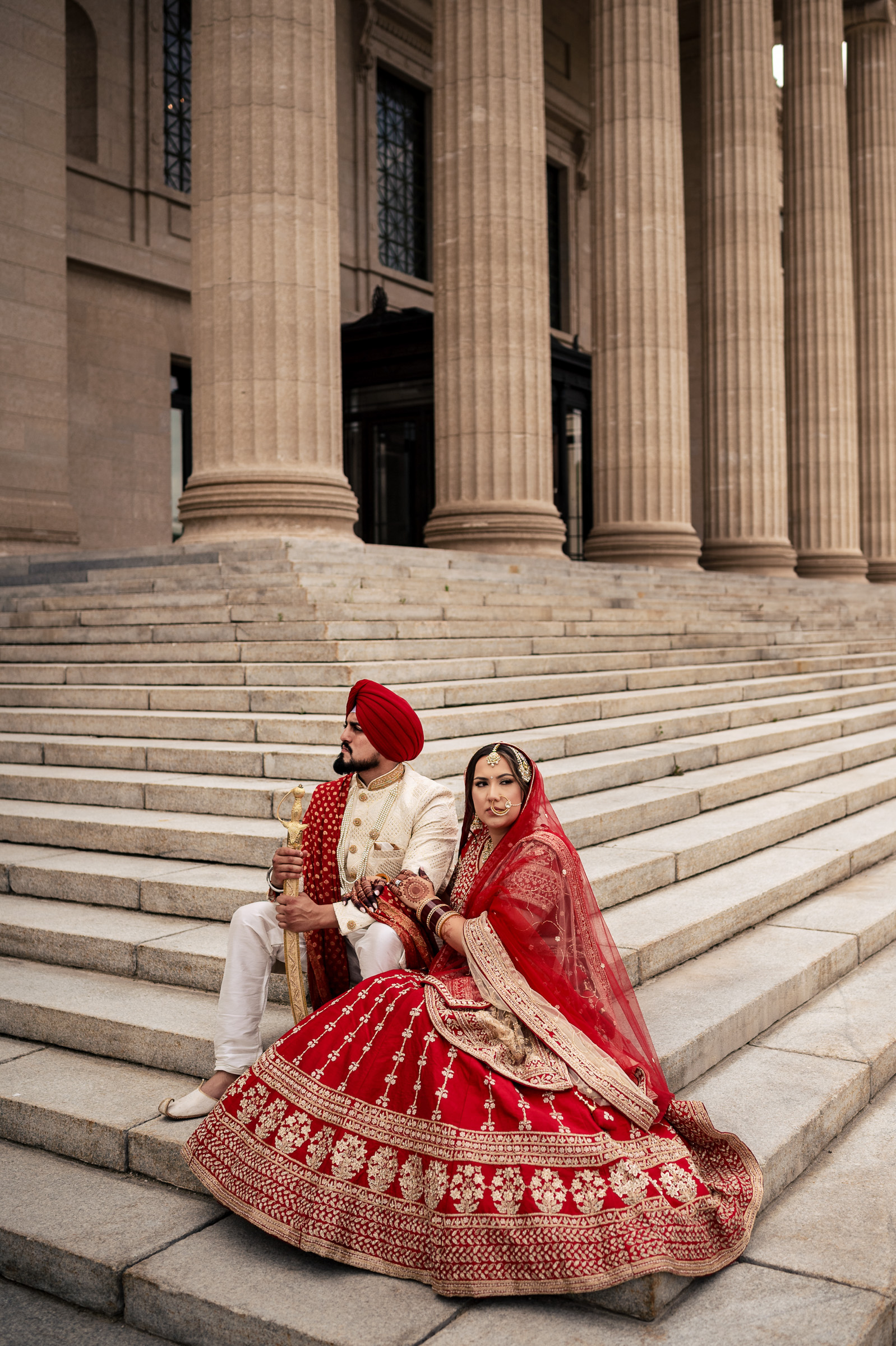 Couple in traditional attire sits on stone steps outside a grand building with columns.