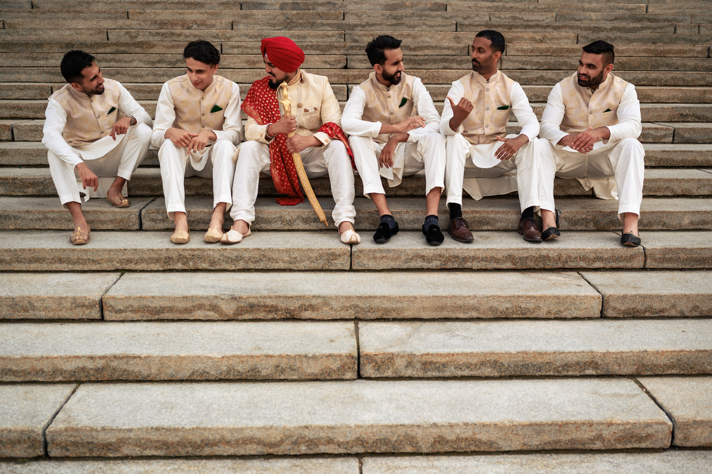 Six men in traditional attire sit on outdoor stone steps, facing each other and talking.