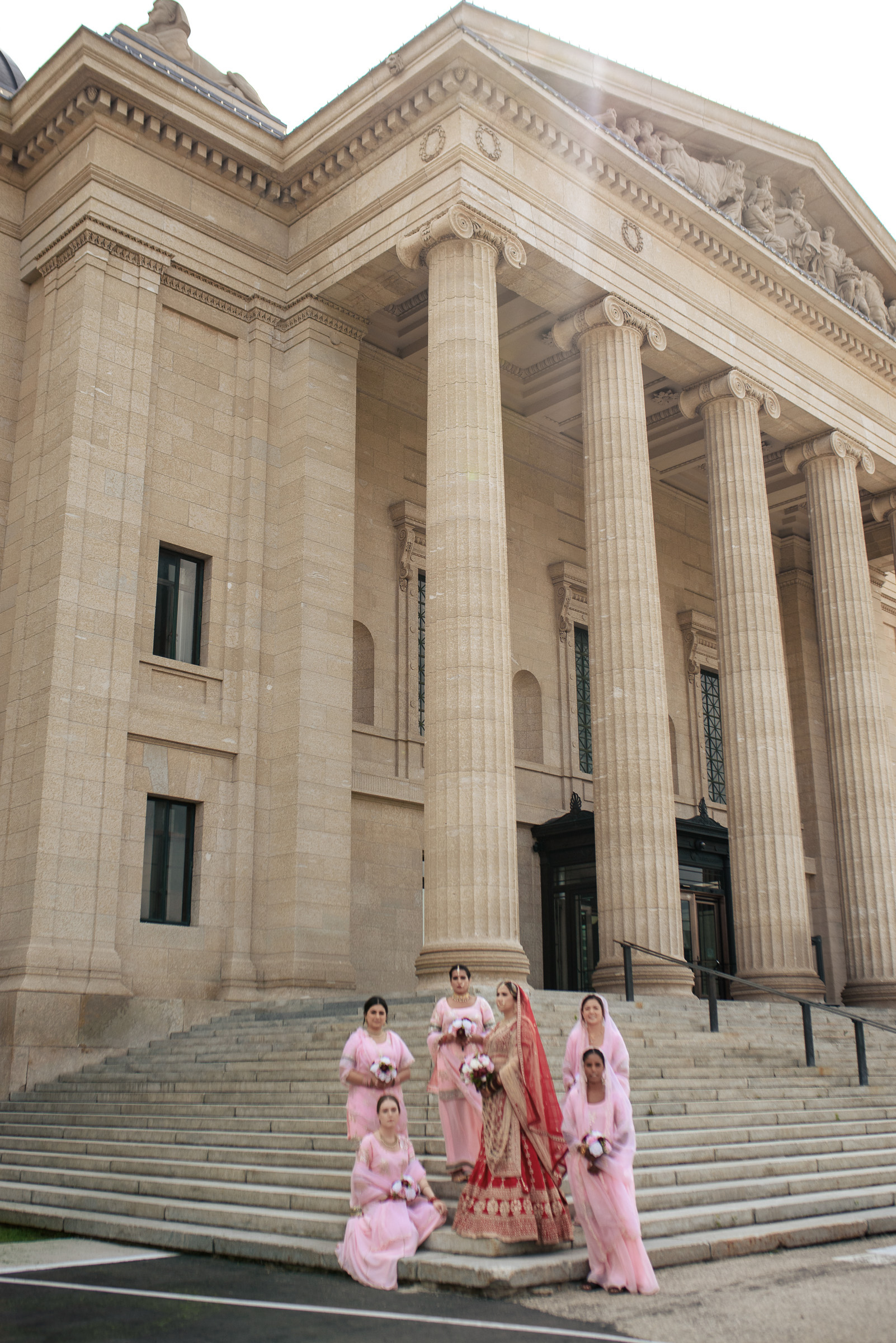 Bride and bridesmaids in pink dresses pose in front of a grand building with columns.