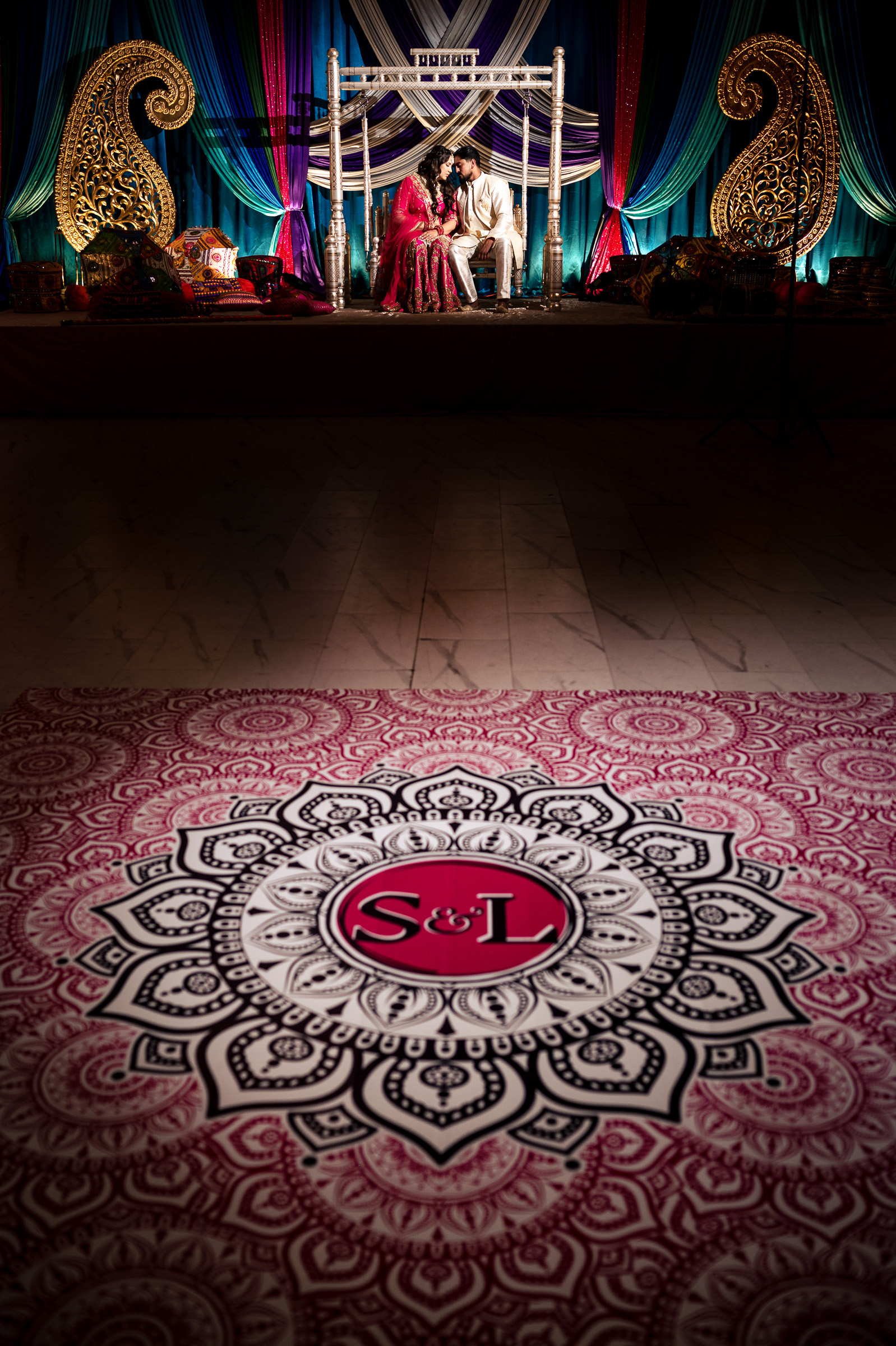 Couple in traditional attire seated on stage with intricate backdrop and decorative floor design.