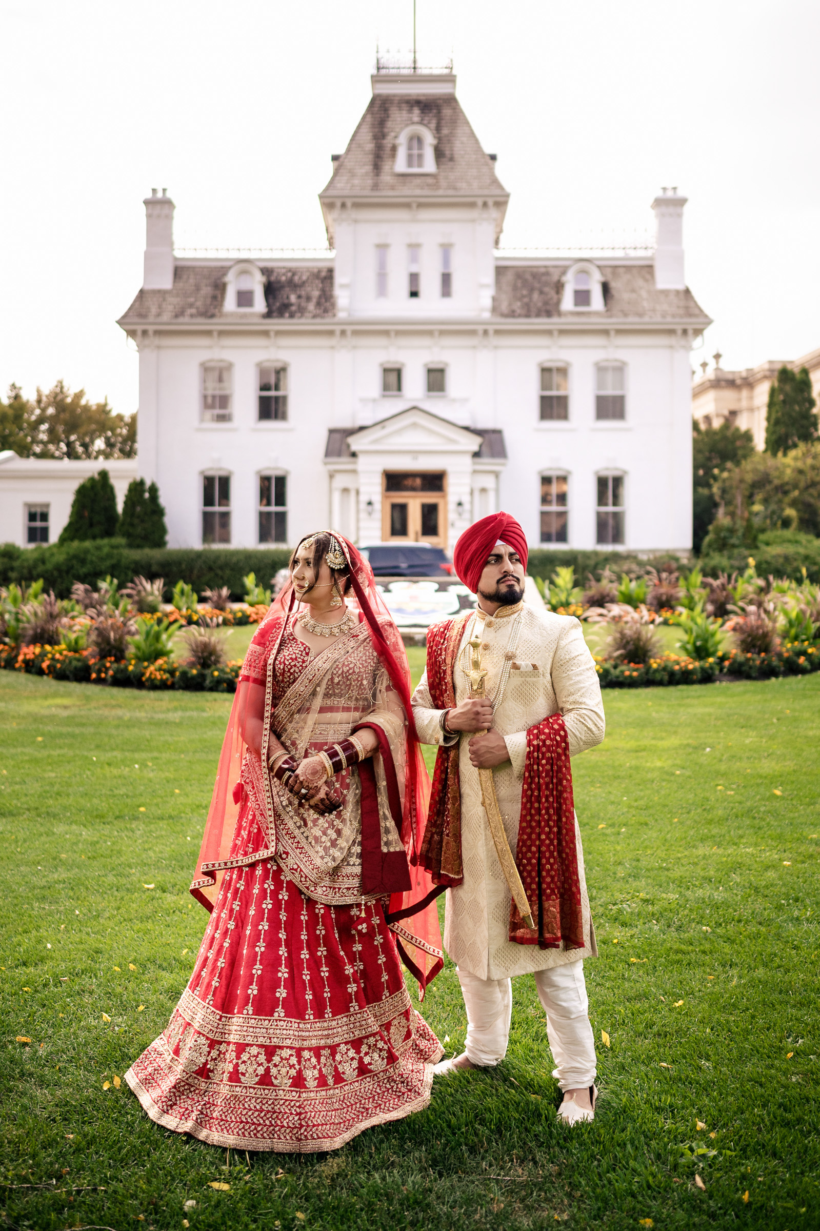 Couple in traditional attire poses in front of a grand white mansion with manicured gardens.