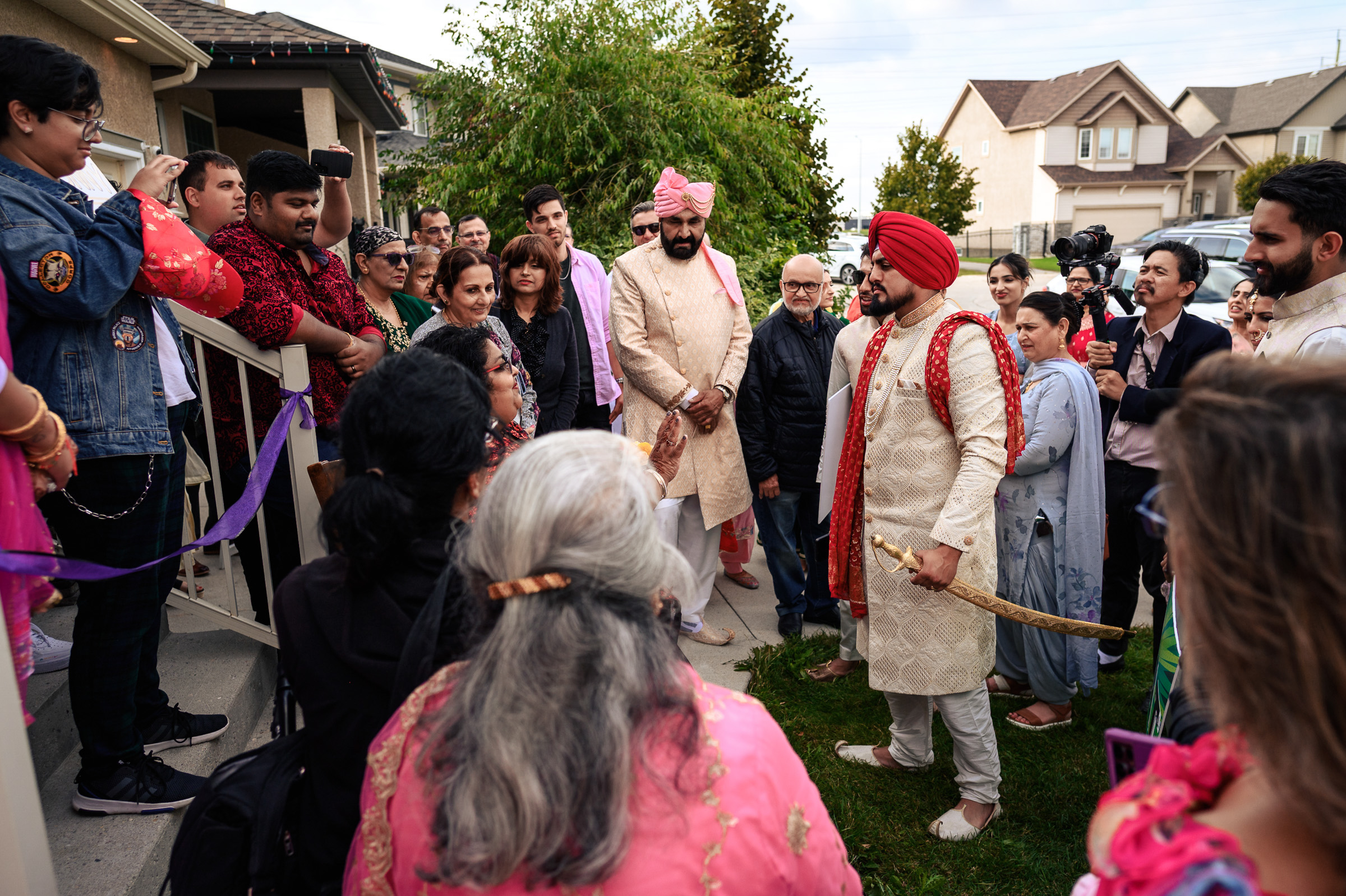 A groom holds a sword outside, surrounded by a group of people in traditional attire.