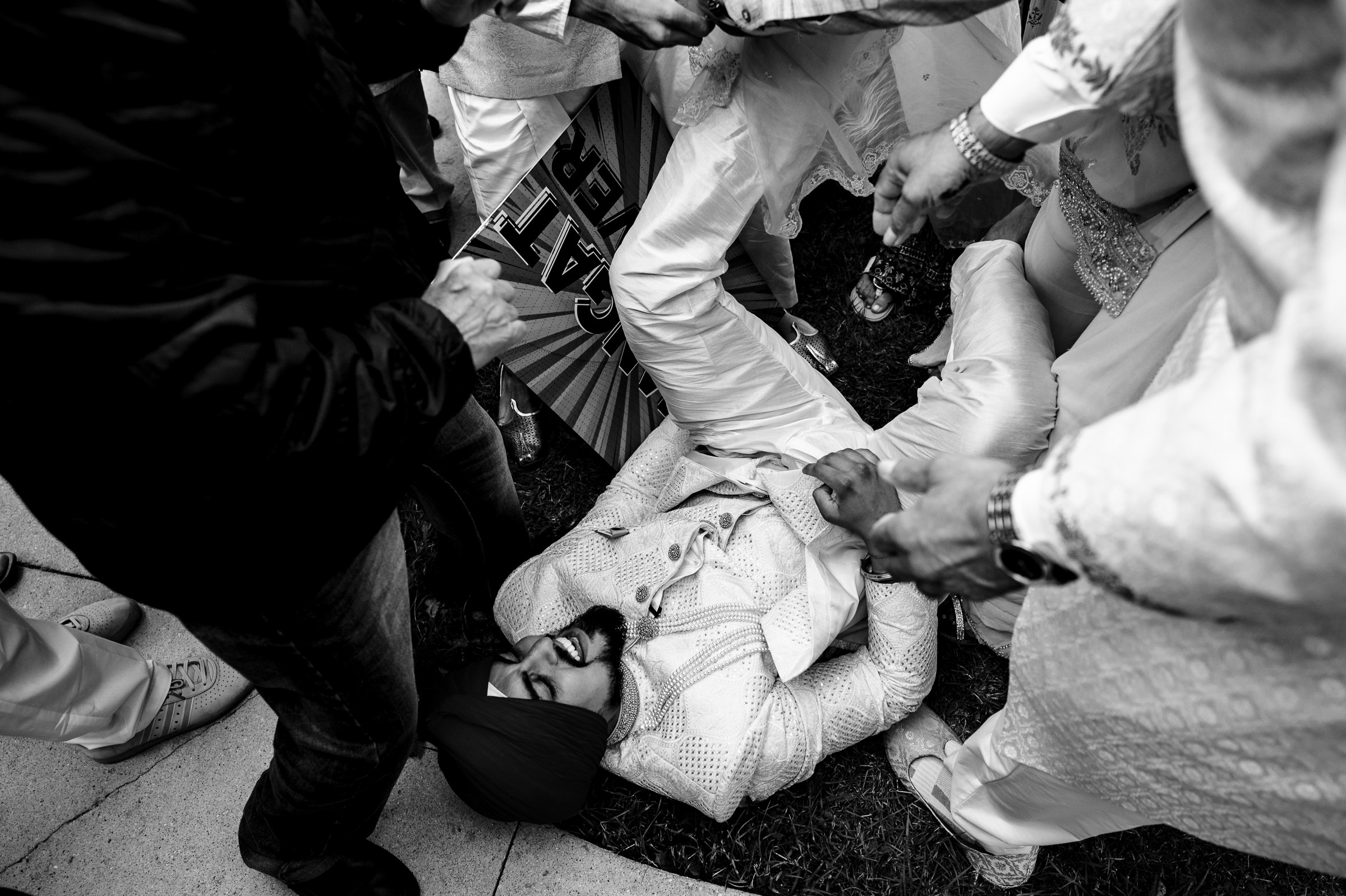 Man in white traditional attire lies on grass, surrounded by people holding his arms.