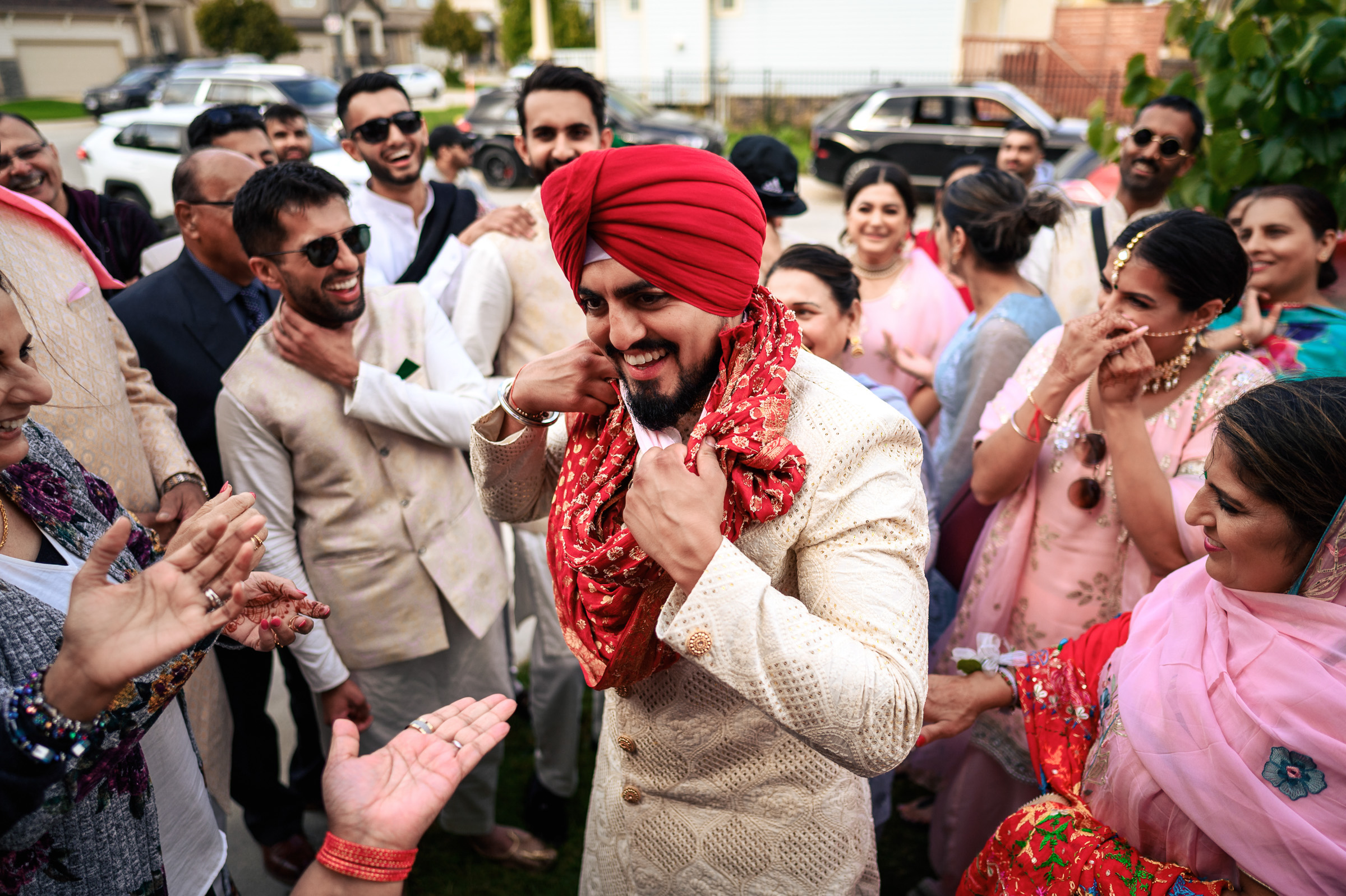A man in a red turban smiles among a cheerful, colorful crowd outdoors.